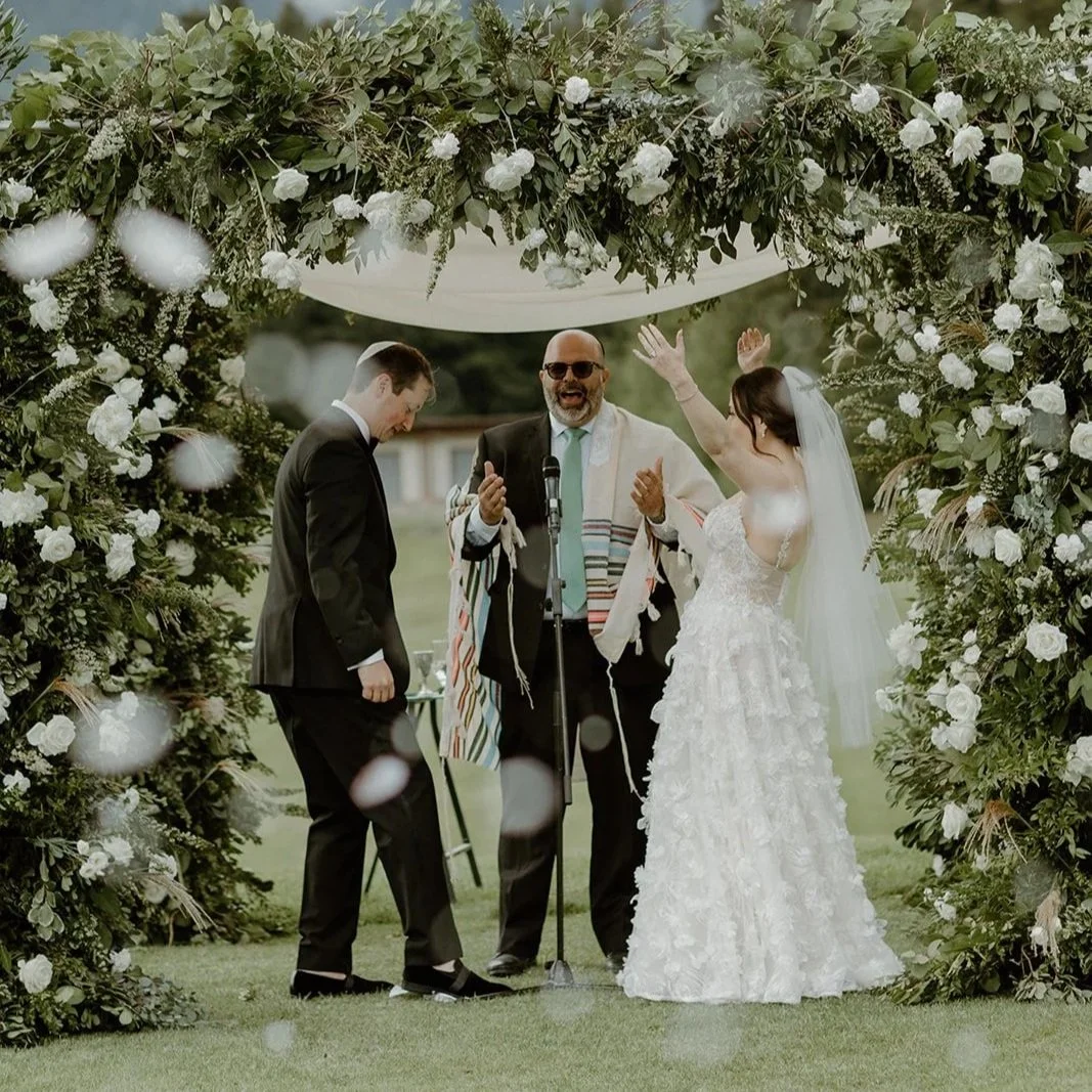 A wedding ceremony outdoors with a bride and groom under a floral arch, with an officiant announcing or speaking, the bride raising her hands, and the groom in a suit smiling, on a grassy area with trees in the background.