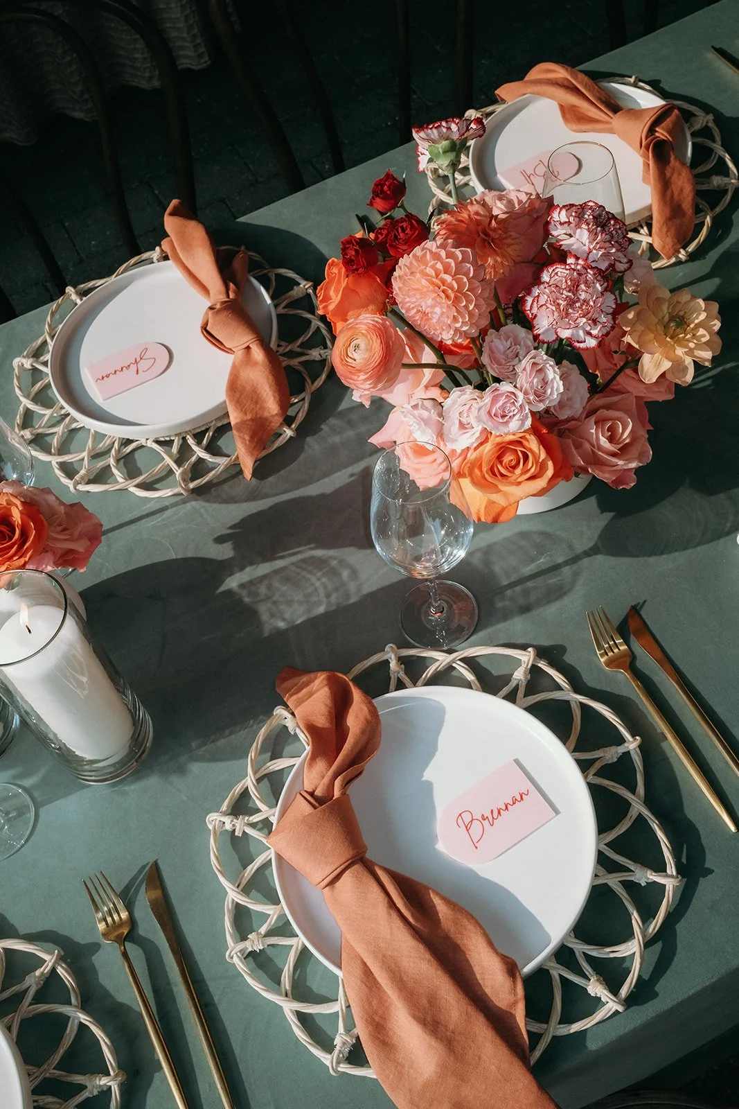Table setting for a formal event with a large pink floral centerpiece, white plates with woven wicker chargers, rust-colored napkins, gold utensils, and place cards with names, including 'Brennan'.