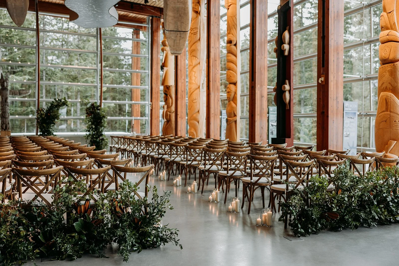 A wooden Whistler wedding ceremony setup with rows of chairs, candles on the floor, and rustic carved wooden columns inside a glass-walled building surrounded by trees.