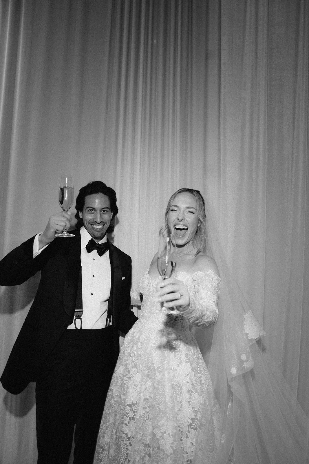 A joyful bride and groom in formal wedding attire celebrating with champagne, standing in front of a curtain, in black and white.