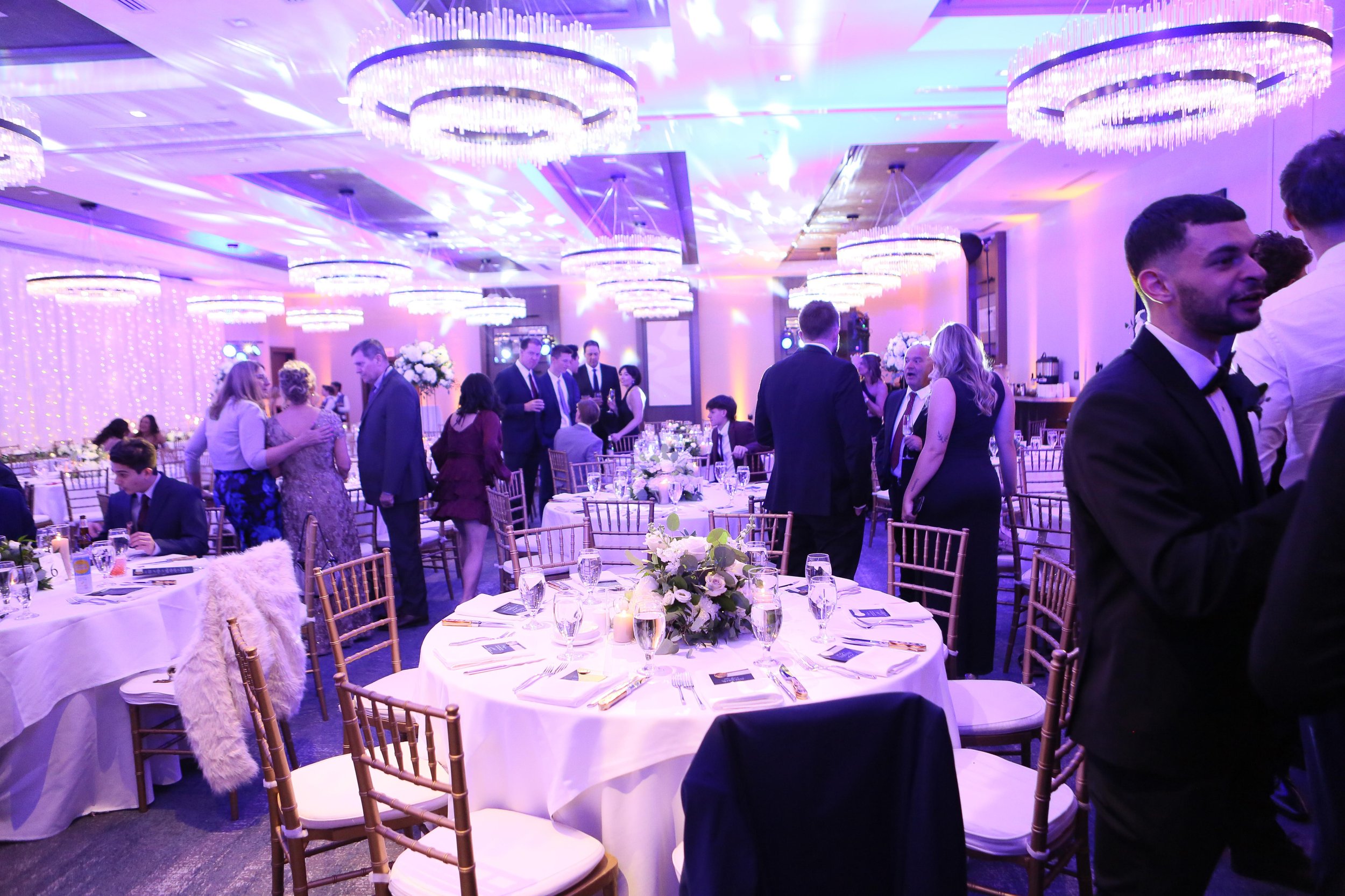 Elegant wedding reception with guests chatting around round tables, adorned with floral centerpieces, under chandeliers and purple lighting.