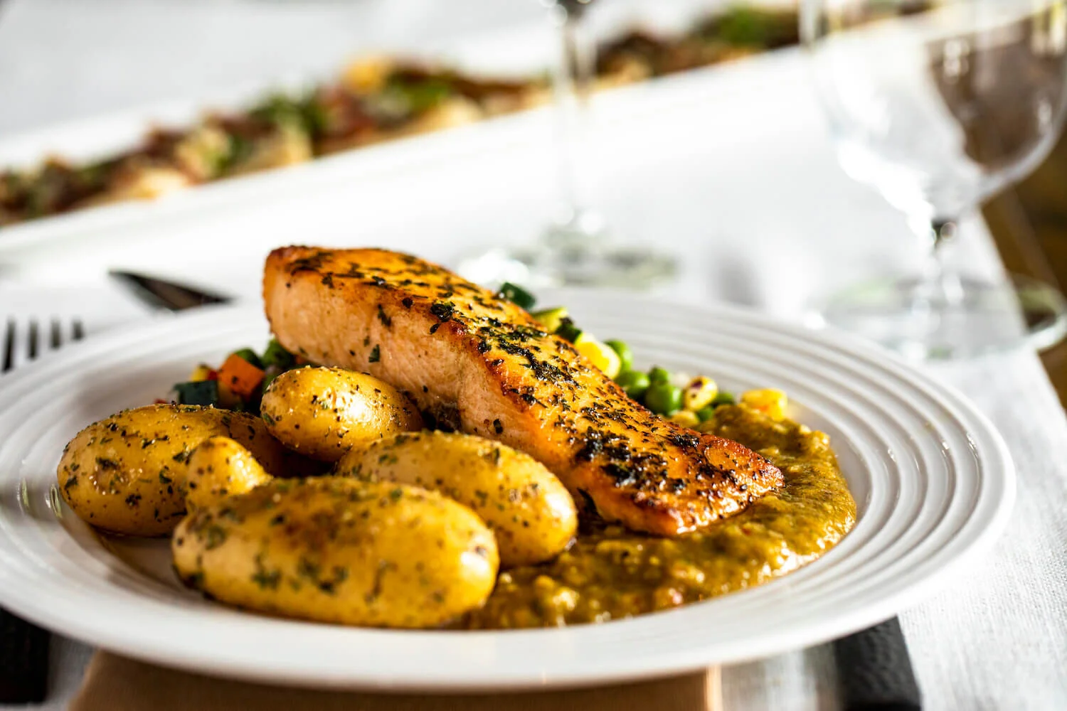 Grilled salmon served with seasoned baby potatoes on a bed of mixed vegetables and puréed sauce on a white plate.