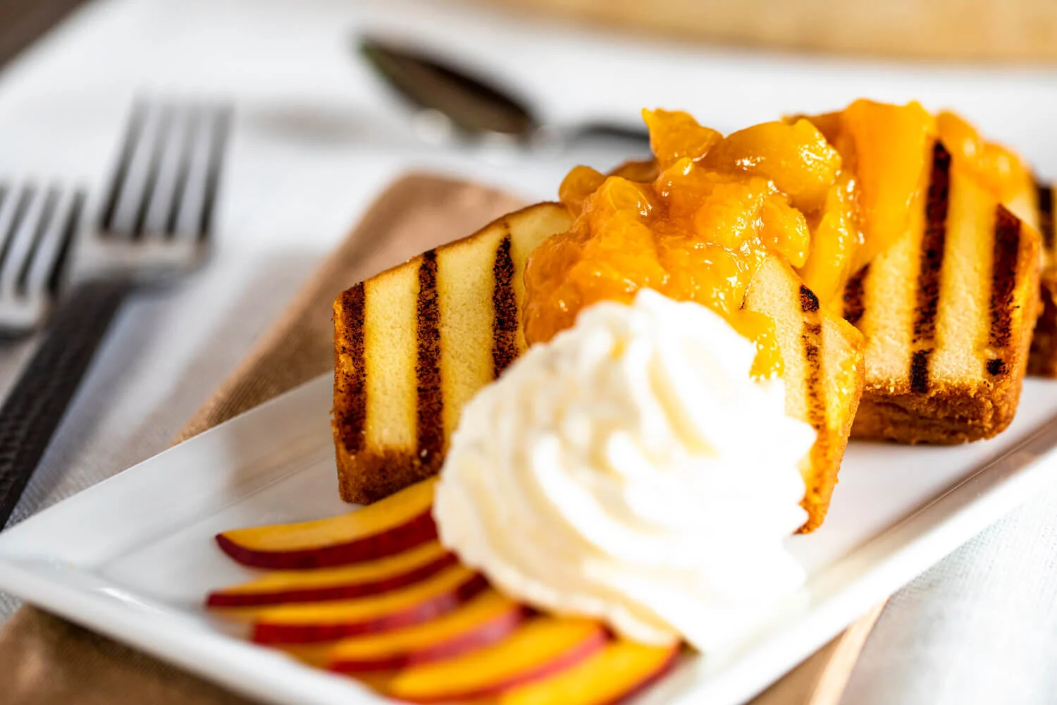 Grilled pound cake with fruit compote, whipped cream, and sliced peaches on a white plate, with forks and a brown napkin nearby.