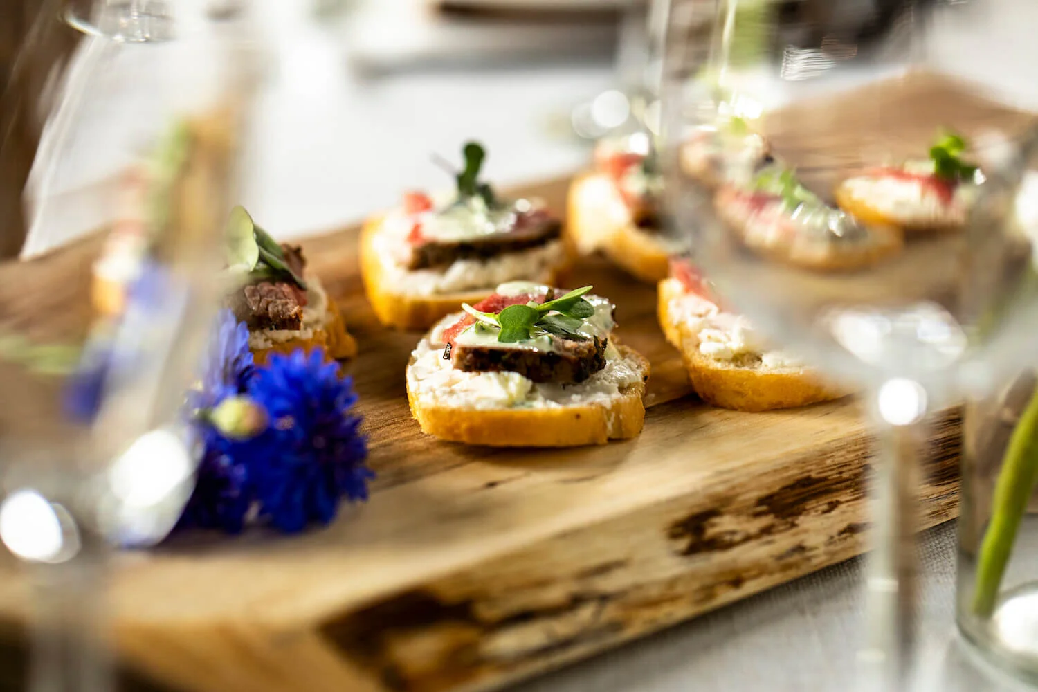 Appetizers on a wooden board with crostini topped with meats, cream, and garnishes; blue flowers and glassware in the foreground.