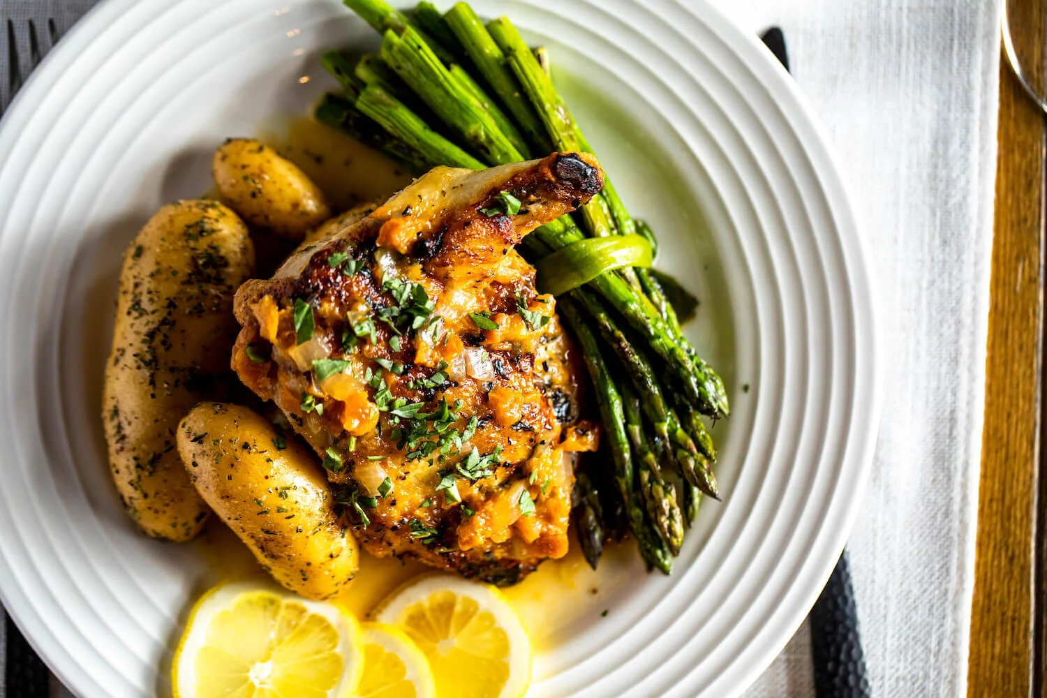 Grilled pork chop with herbs, serving of fingerling potatoes with parsley, roasted asparagus, and lemon slices on a white plate.