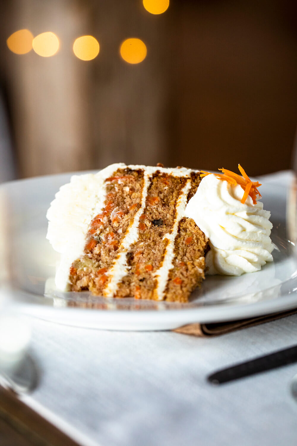 A slice of carrot cake with cream cheese frosting on a plate, garnished with whipped cream and carrot shavings, set on a table with blurred lights in the background.