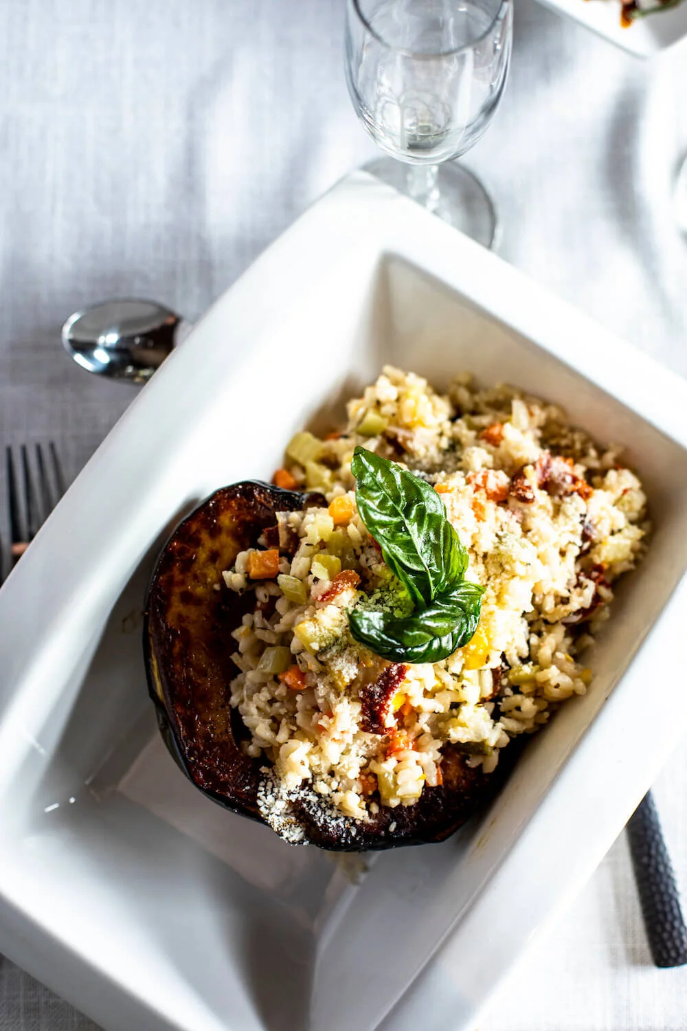 Stuffed acorn squash with rice and vegetables, garnished with a basil leaf, served in a white rectangular plate on a table setting with silverware and a wine glass.