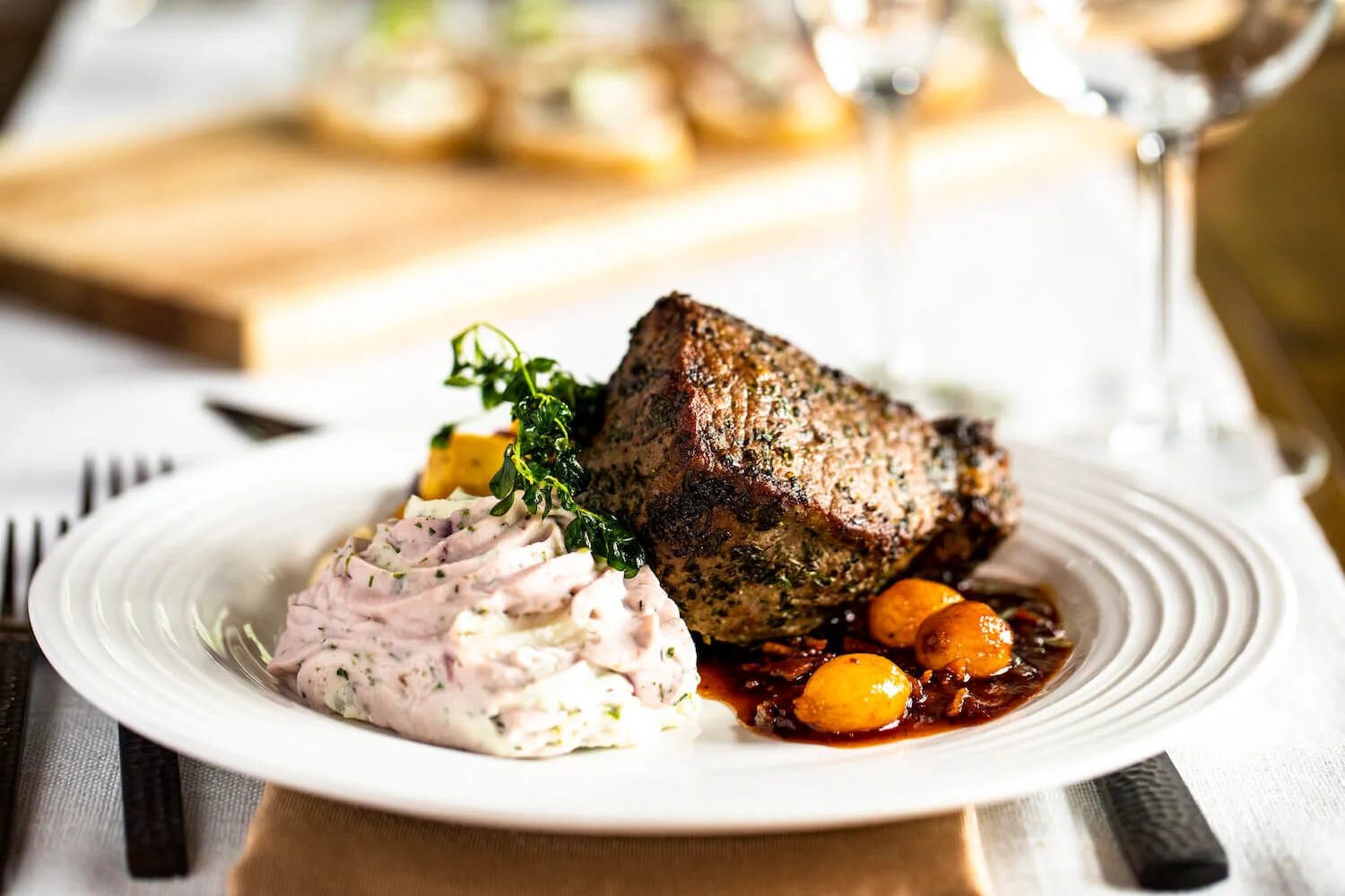 Seared steak with mashed potatoes, roasted tomatoes, and herbs on a white plate, set on a white tablecloth.