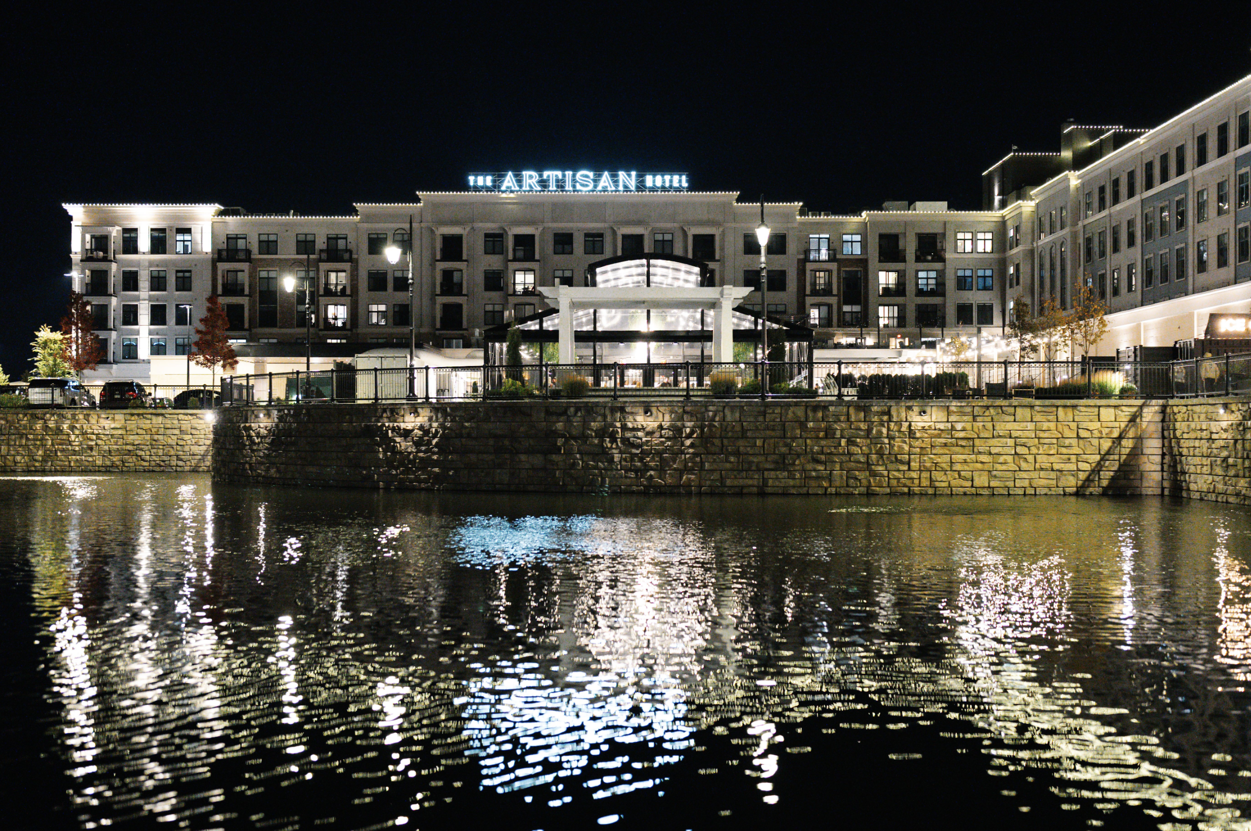 Nighttime view of The Artisan Hotel with illuminated windows and reflections on water.