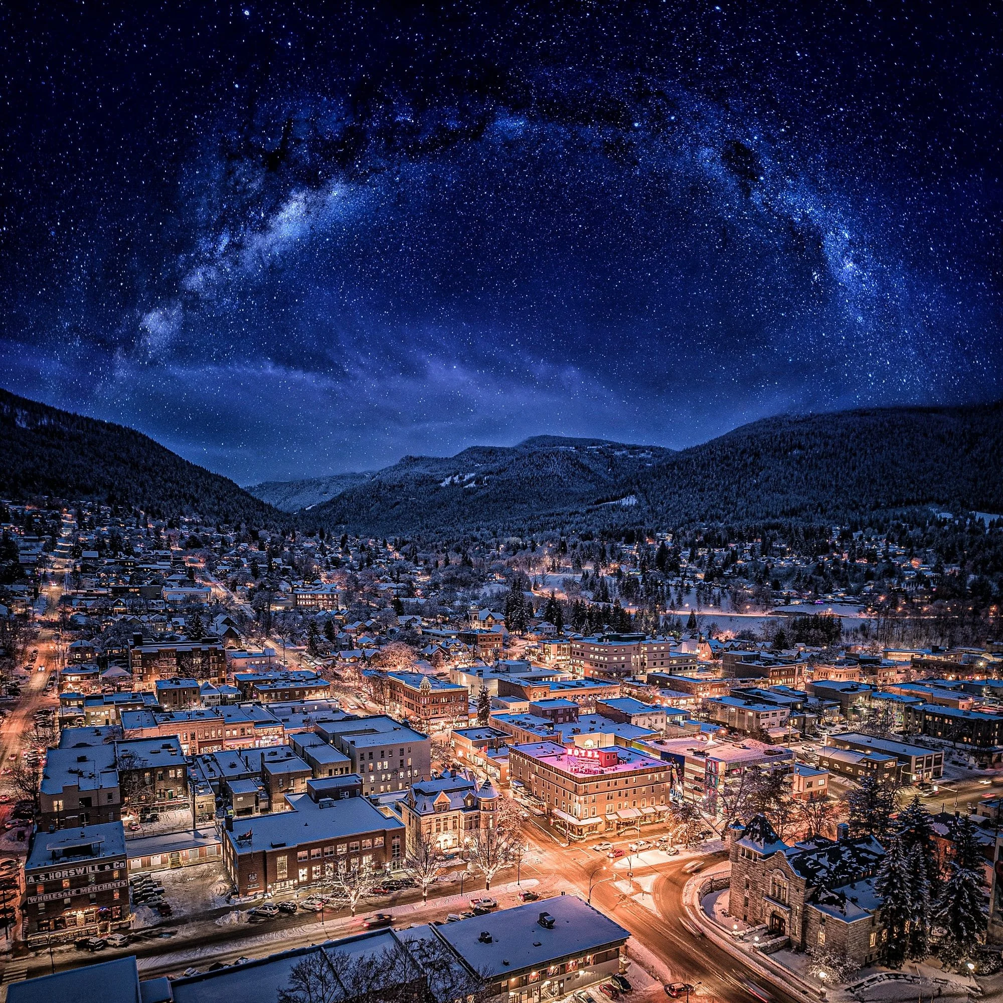 Nighttime view of a snow-covered town with colorful lights, surrounded by mountains and a starry sky with the Milky Way galaxy.