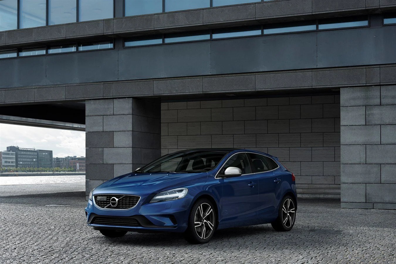 A blue Volvo hatchback parked on a cobblestone road in front of a modern building with large windows and gray stone walls.