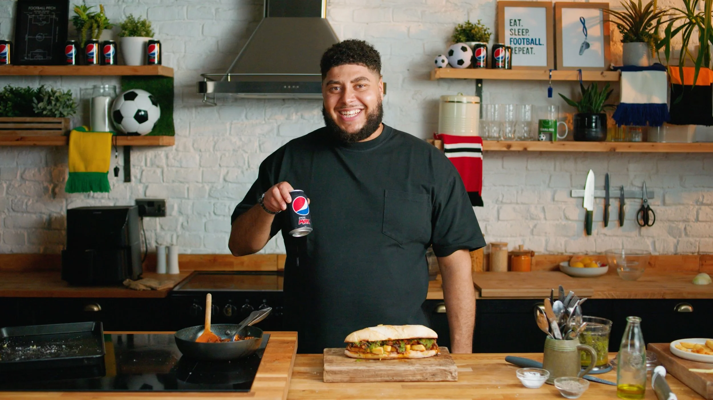 A man with a beard and curly hair smiling and holding a Pepsi can in a kitchen. There is a sandwich on a wooden board on the counter in front of him. The kitchen has white brick walls, wooden shelves with plants, soccer balls, and kitchen utensils, and a framed sign that says, "Eat. Sleep. Football. Repeat."