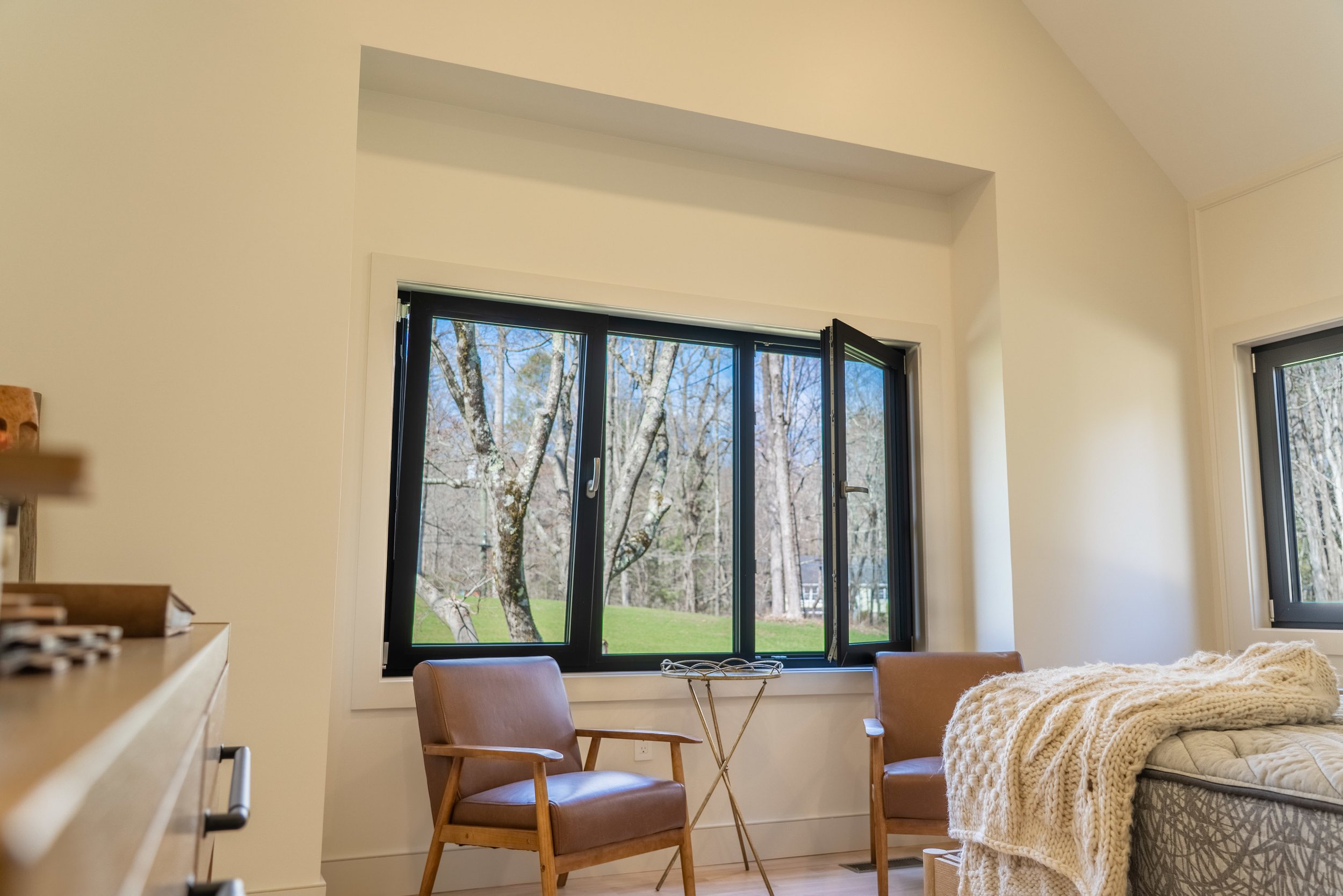 Modern living room with two brown leather chairs, a small round table, and large windows showing a scenic outdoor view. The windows are framed with black trim. There is a bed with a beige knitted throw blanket in the foreground.
