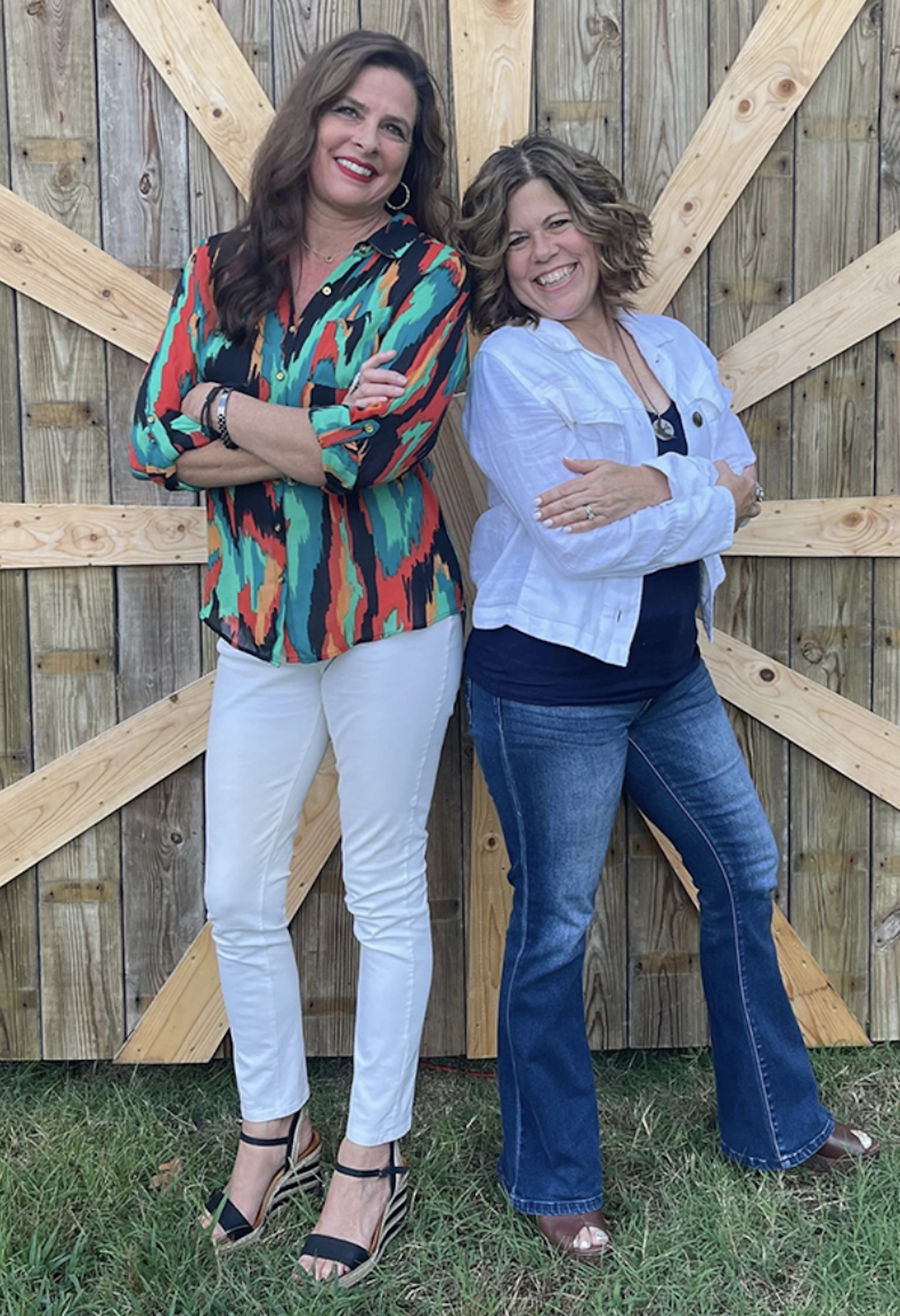 Two women standing side by side outdoors in front of a wooden fence, smiling and crossing their arms.