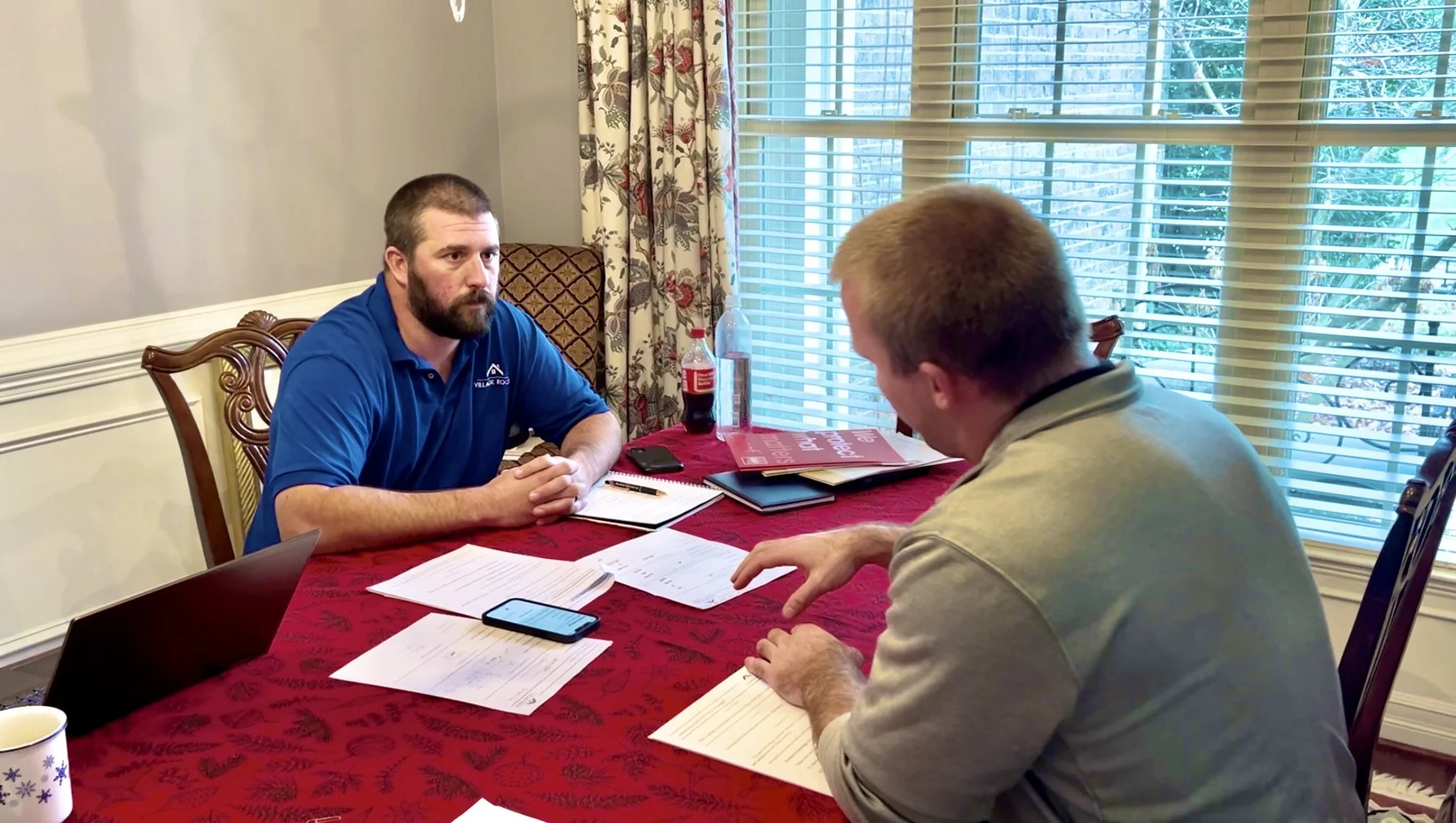 Two men sitting at a table in a sunlit room, engaged in a discussion with documents, notebooks, a phone, and a laptop on the table, with windows and floral curtains in the background.