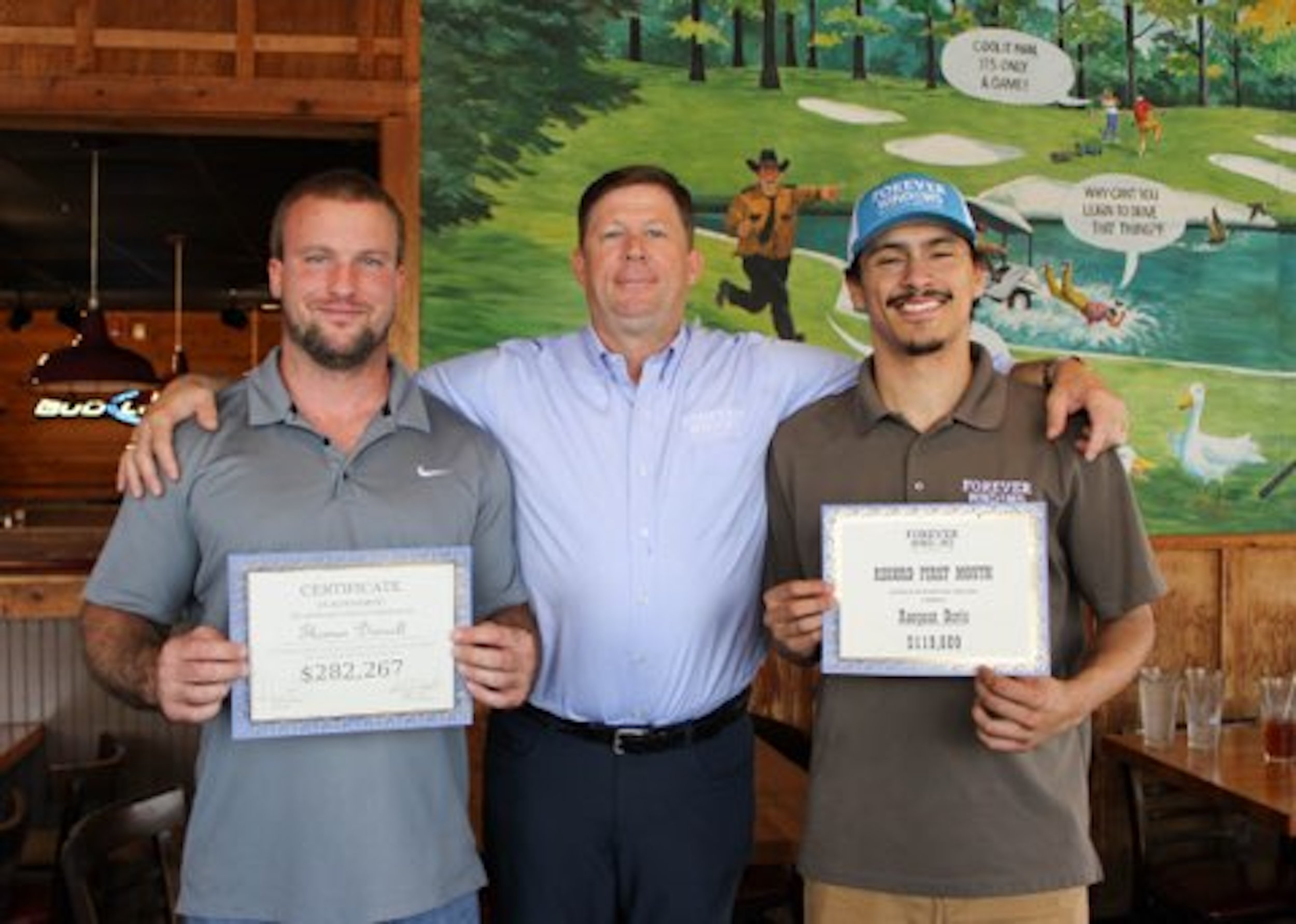 Three men standing inside a restaurant, with two of them holding certificates. The man in the middle has his arms around the other two. Behind them is a colorful mural depicting a golf course scene.