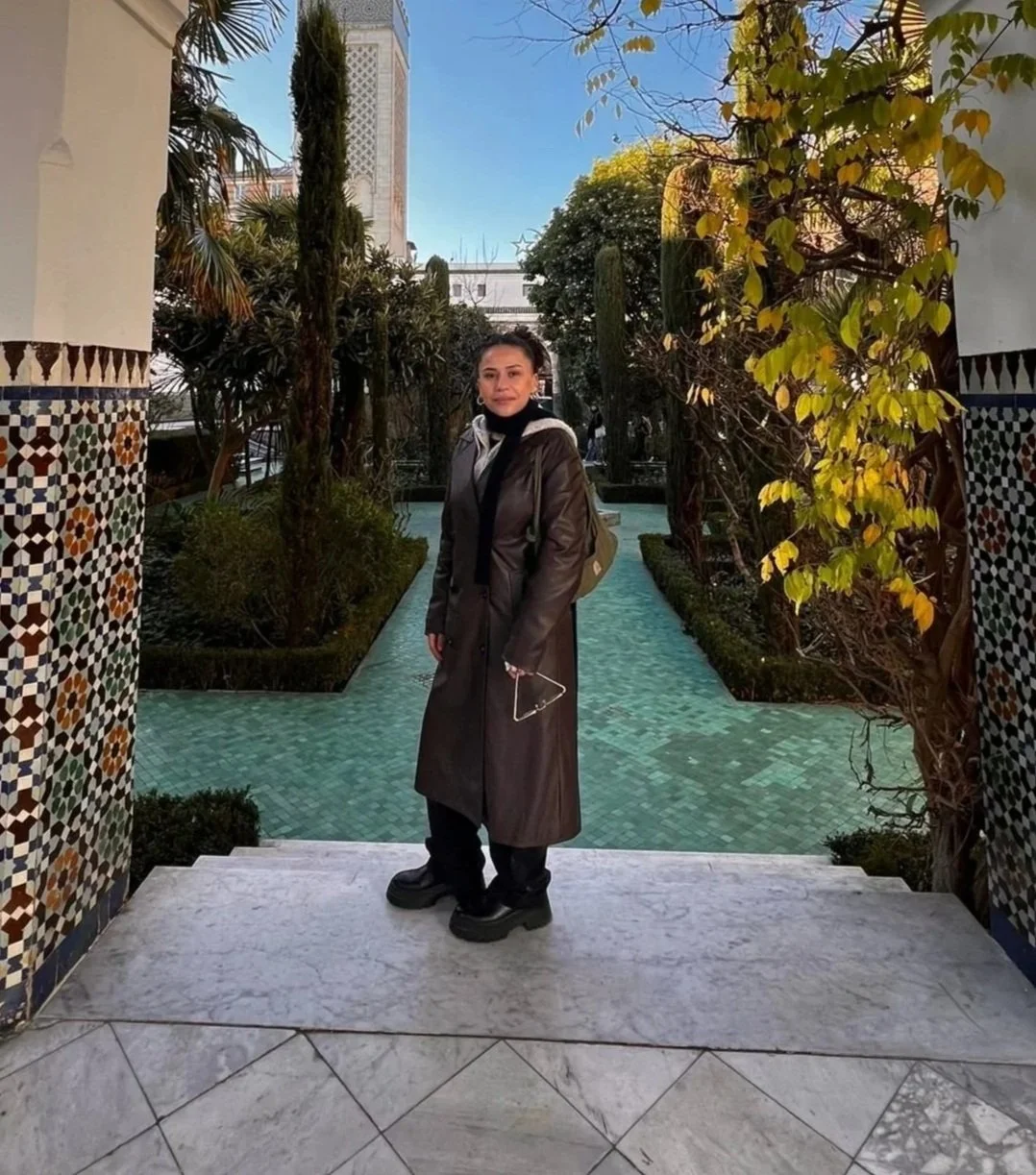 A woman in a black coat and boots standing on a marble step outdoors, holding a phone, with lush garden and cityscape in the background.
