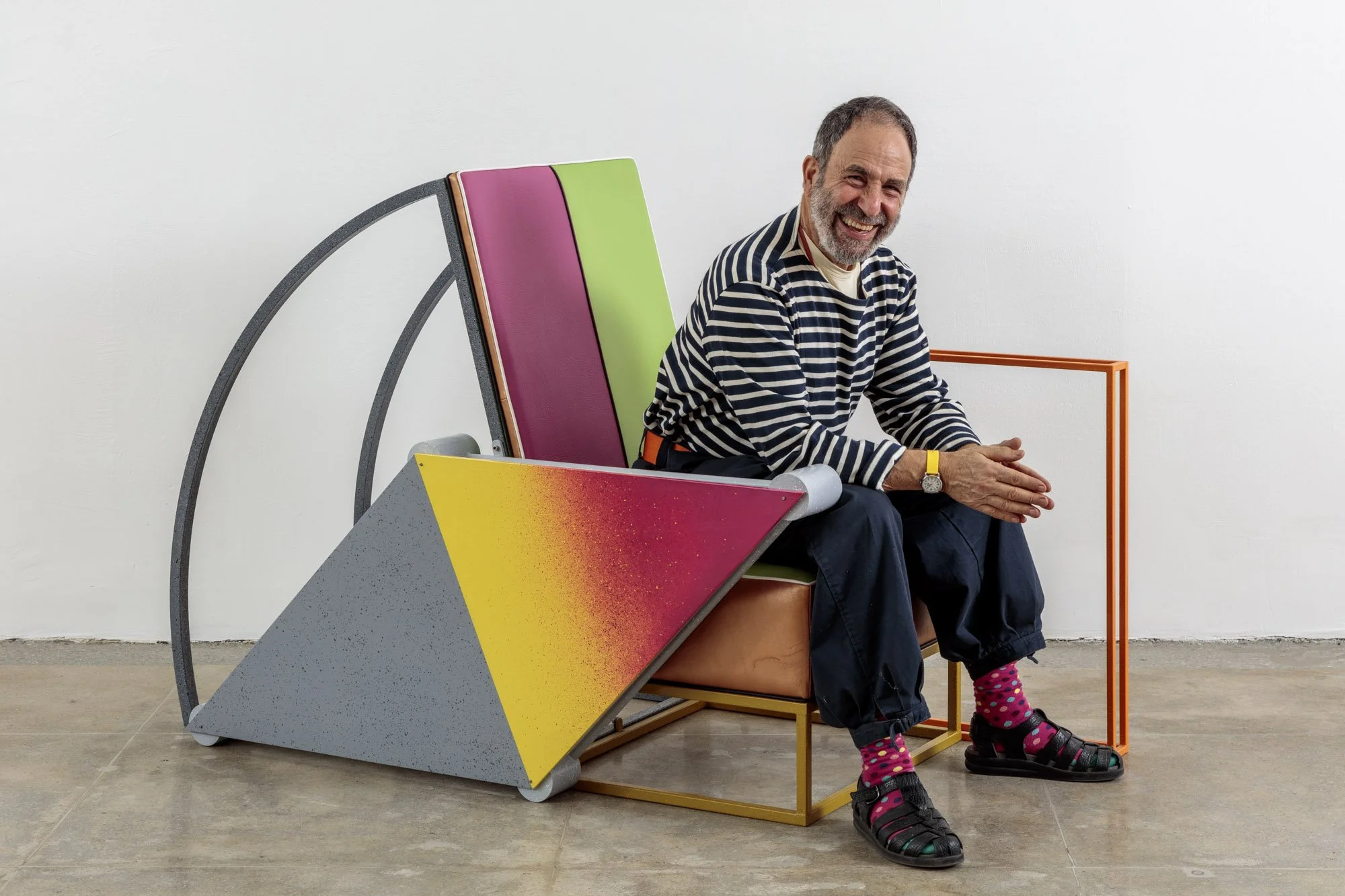 Artist Peter Shire is wearing a striped shirt, dark pants, and colorful socks sits on a modern chair with geometric design elements in a white room, and is smiling at the camera.