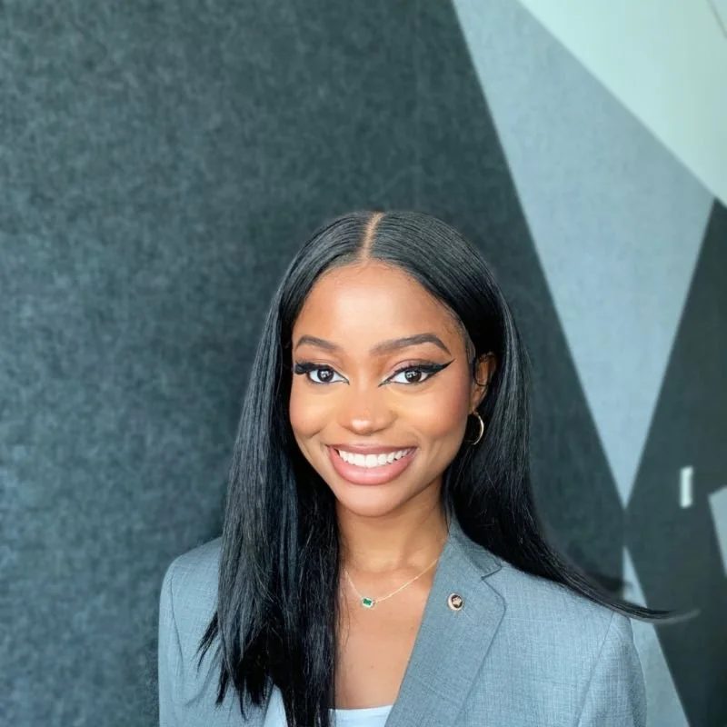 A smiling young woman with long black hair, wearing a gray blazer and jewelry, standing against a dark, textured background