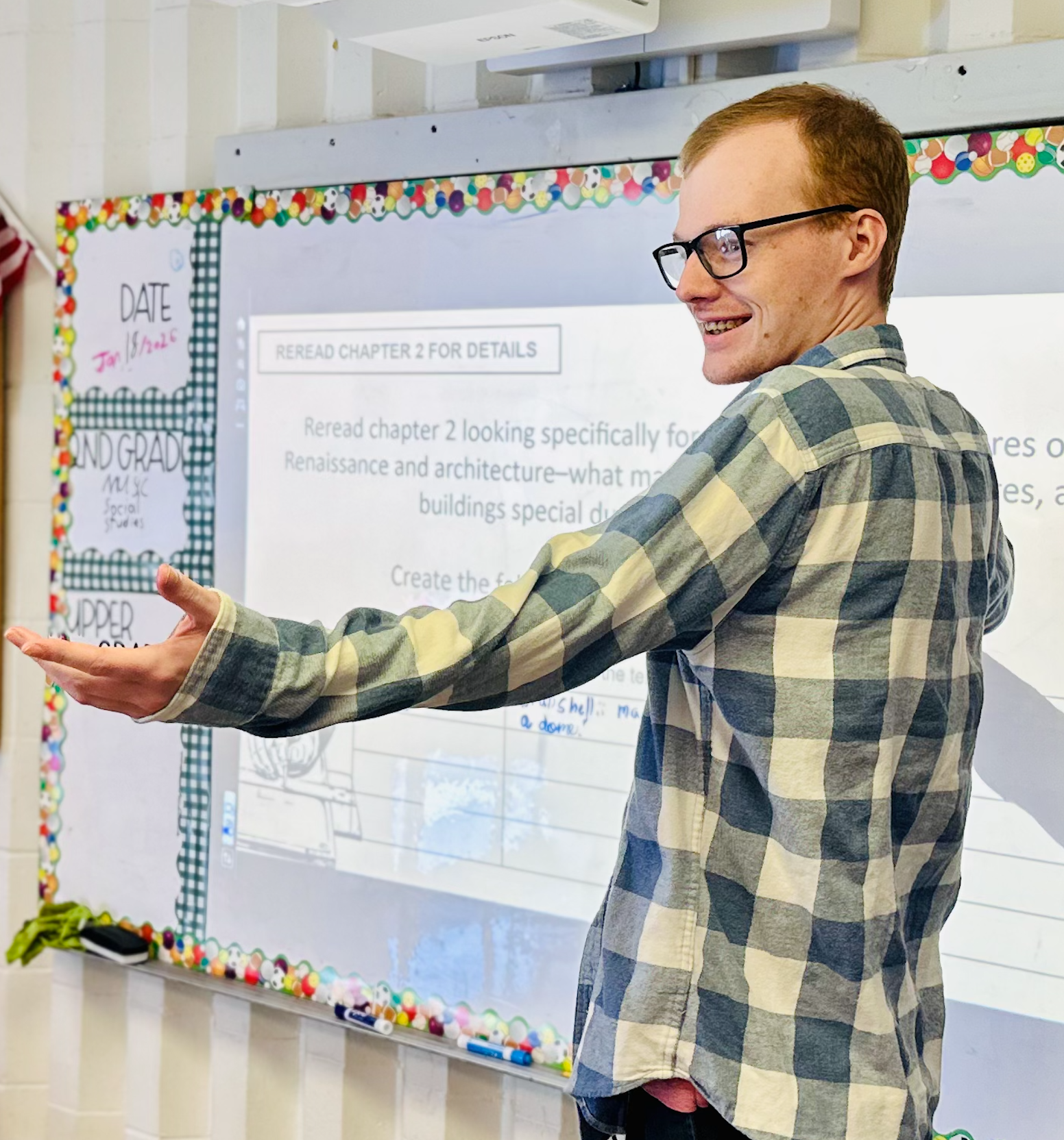 Man with glasses smiling in front of whiteboard and gesturing welcomingly
