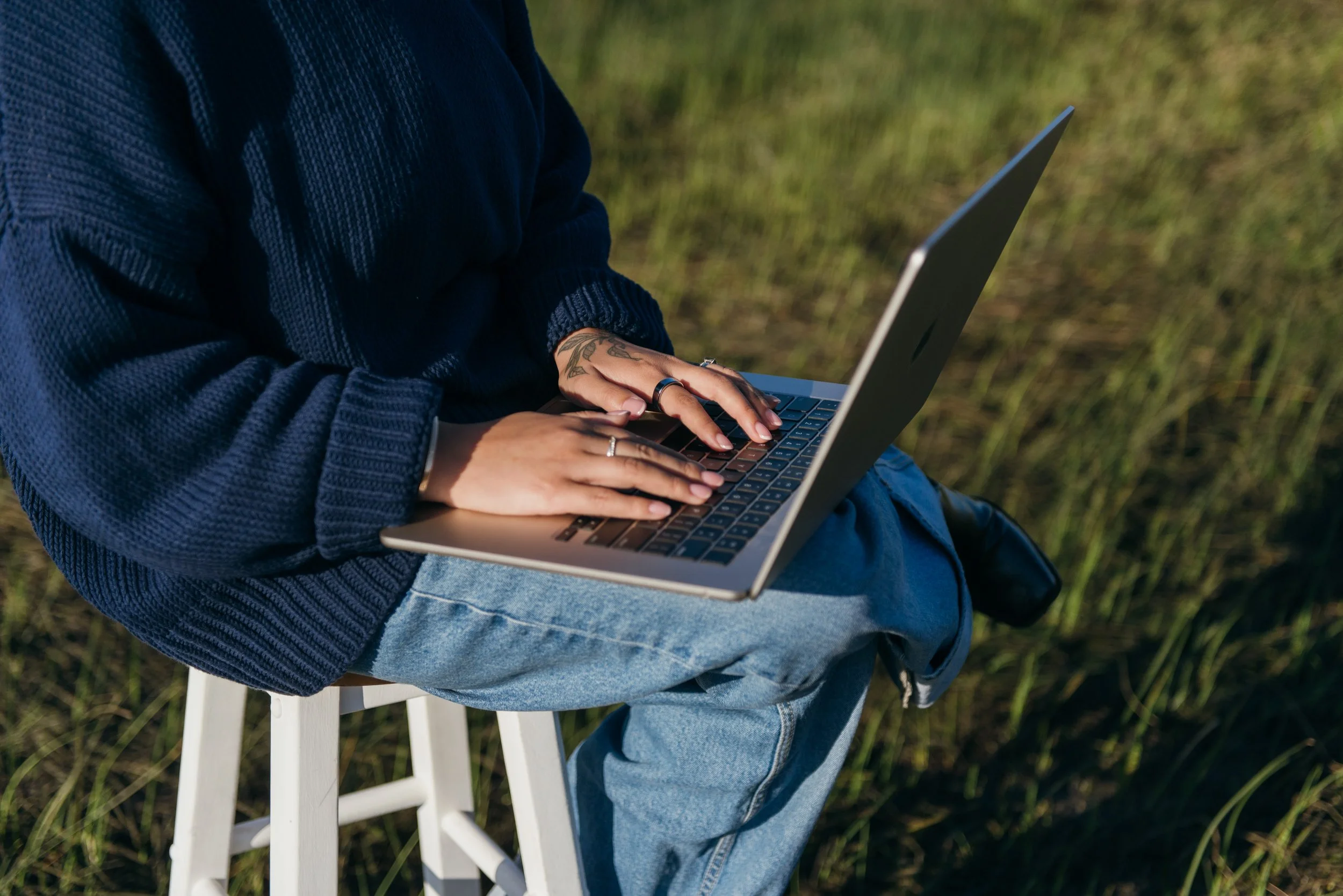 Person sitting on a white stool outdoors, typing on a silver laptop, wearing a navy sweater and light blue jeans, with green grass in the background.