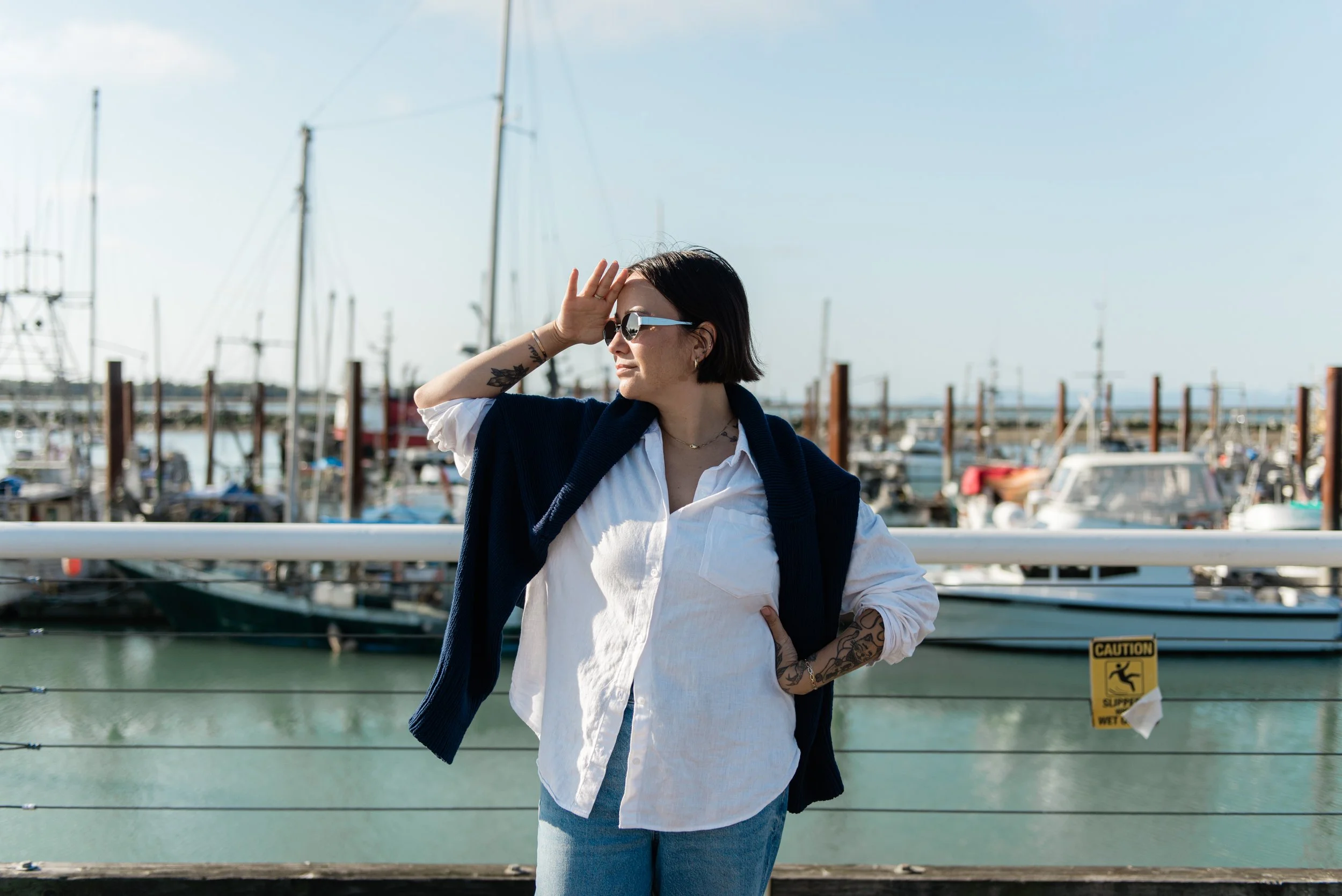 Woman with sunglasses and tattoos gazing out at a marina of sailboats and boats on a sunny day.