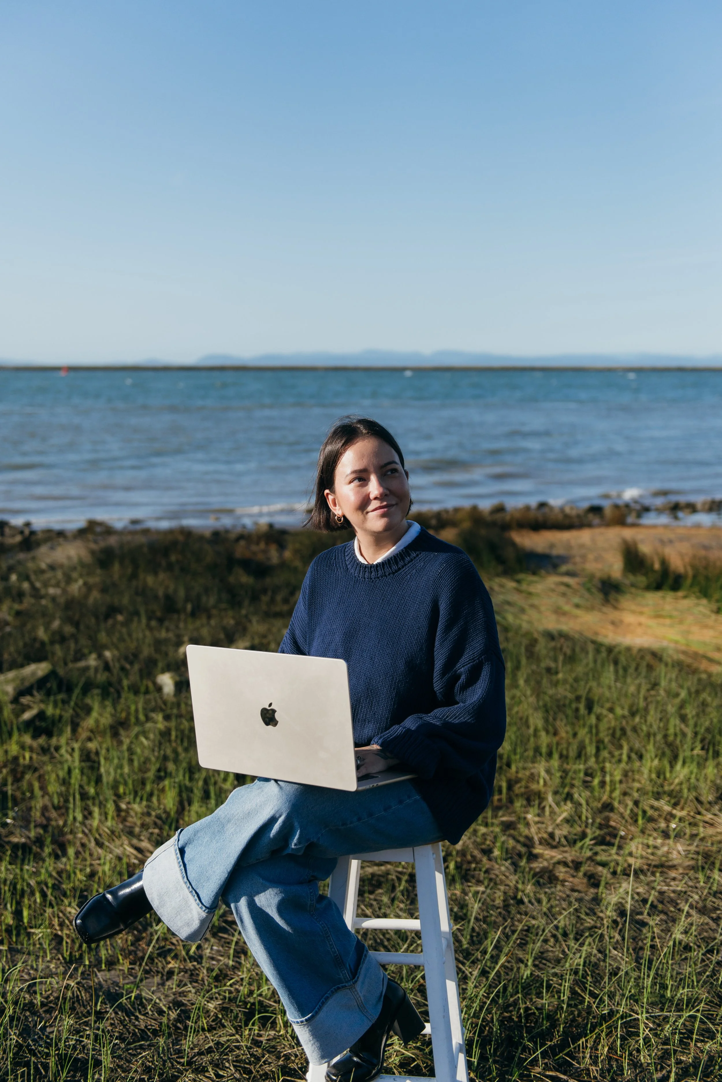 A woman sitting on a white stool outdoors near a body of water, holding a silver MacBook laptop on her lap, with the water and a clear blue sky in the background.