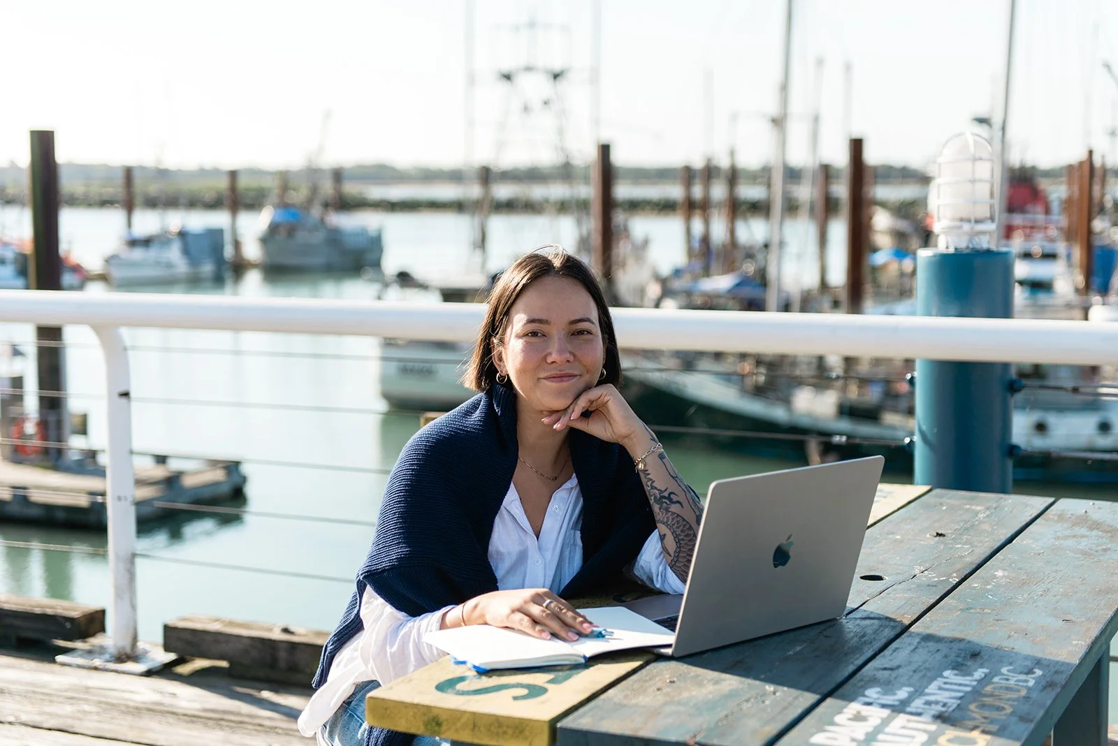 A woman sitting at a table near a marina with boats docked in the background. She has short dark hair, wears a white shirt with a navy sweater draped over her shoulders, and has a tattoo on her arm. She is smiling slightly, resting her chin on her hand, with a silver laptop and a notebook in front of her.