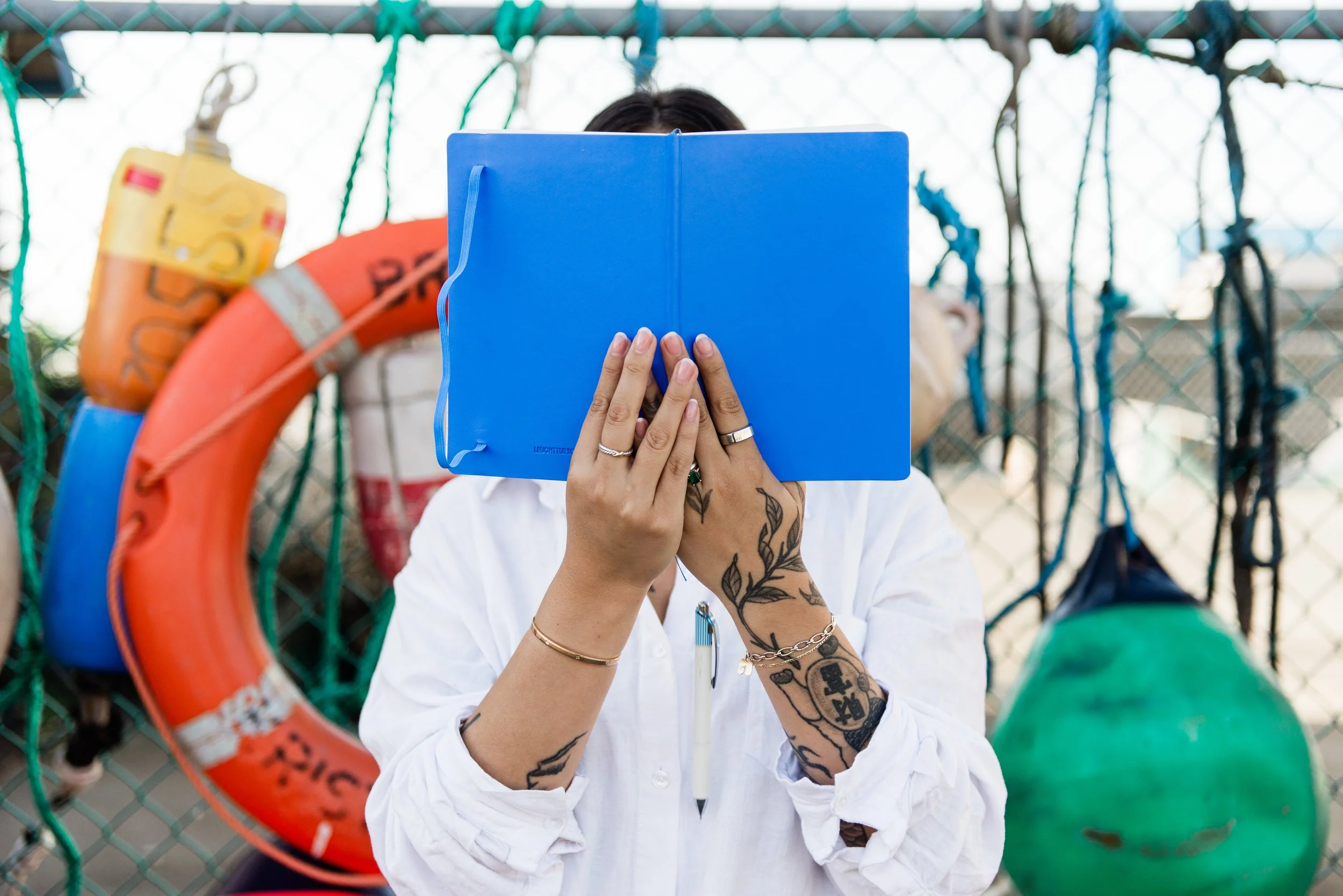 Person holding a blue folder in front of their face, wearing rings, bracelets, and tattoos, standing near a fence with life preservers and equipment hanging on it.