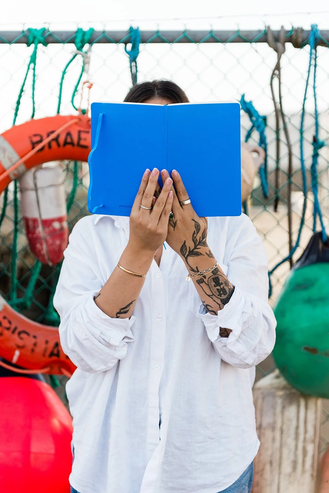 Person with tattoos covering their arms reading a blue booklet outdoors near a chain-link fence, with life preservers and buoys hanging in the background.
