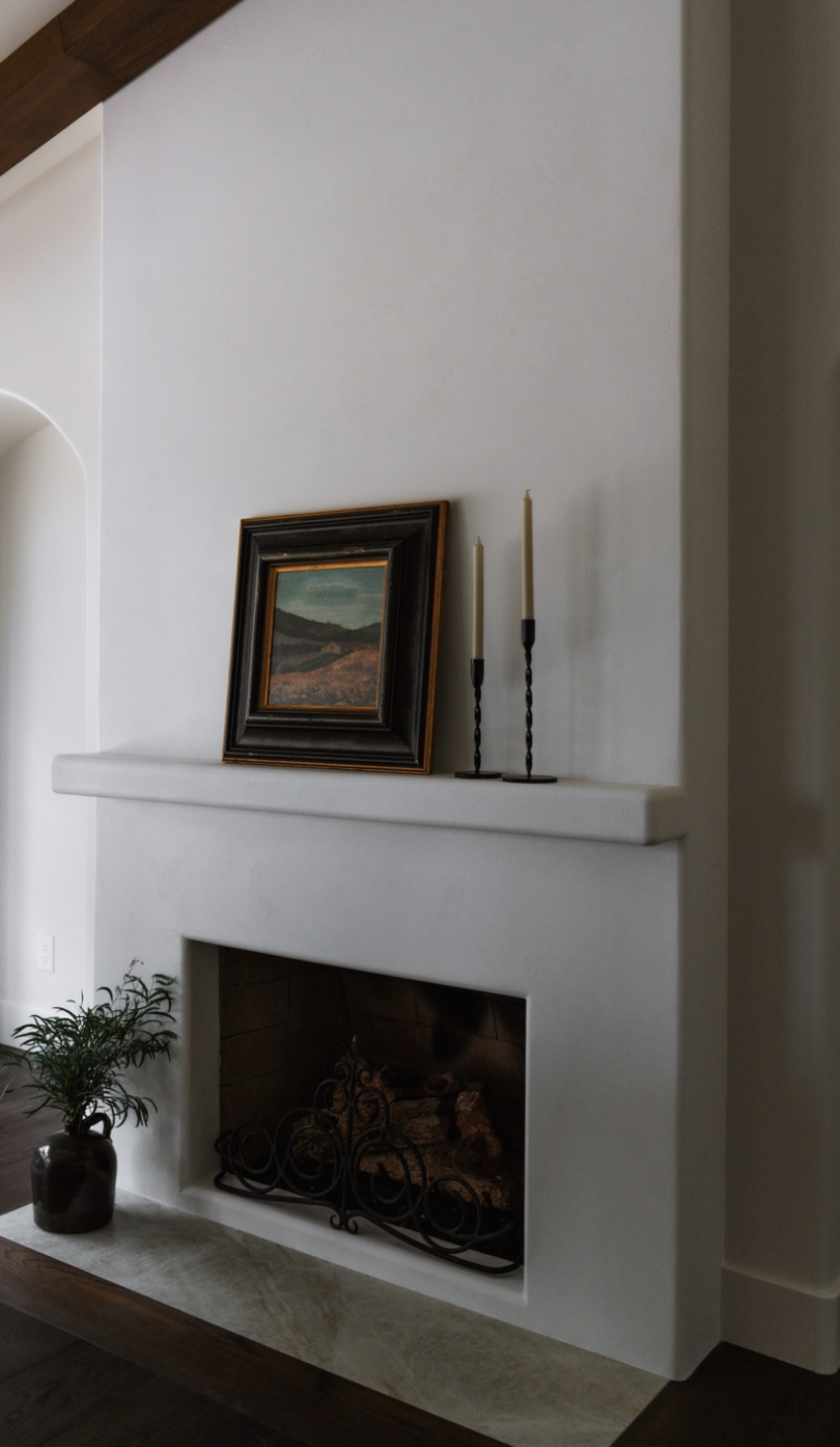 White fireplace with a small landscape painting in a black frame and two tall candlesticks on the mantel. A potted plant is on the floor beside it.