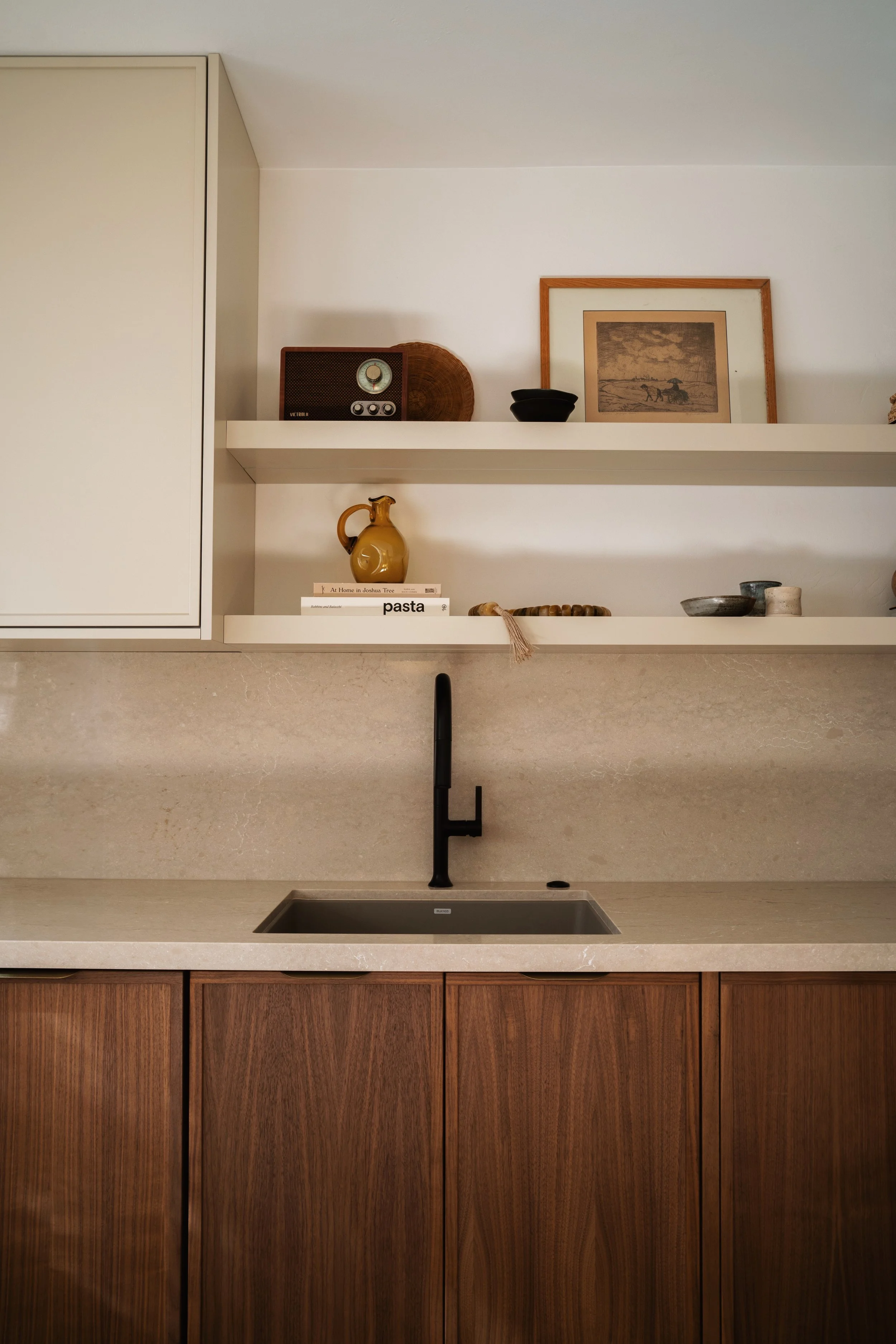 Kitchen countertop with wood cabinets, a black sink, and a beige marble surface. Open shelving above with decorative items including a vintage radio, a framed artwork, a yellow pitcher, and various bowls and books.