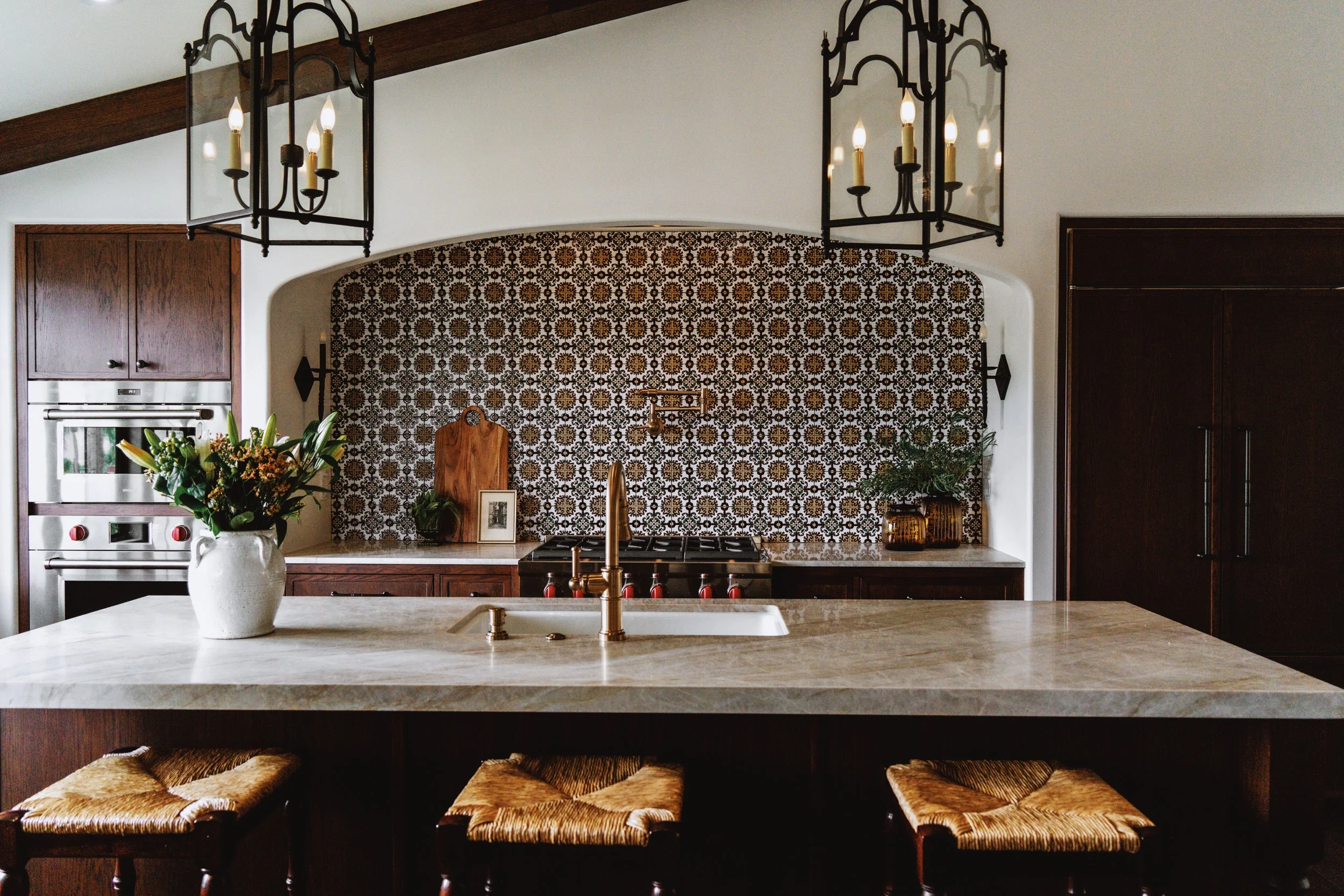 Modern kitchen with black wrought iron chandeliers, patterned backsplash, wooden cabinets, and a white marble island with a vase of flowers and bar stools with woven seats.