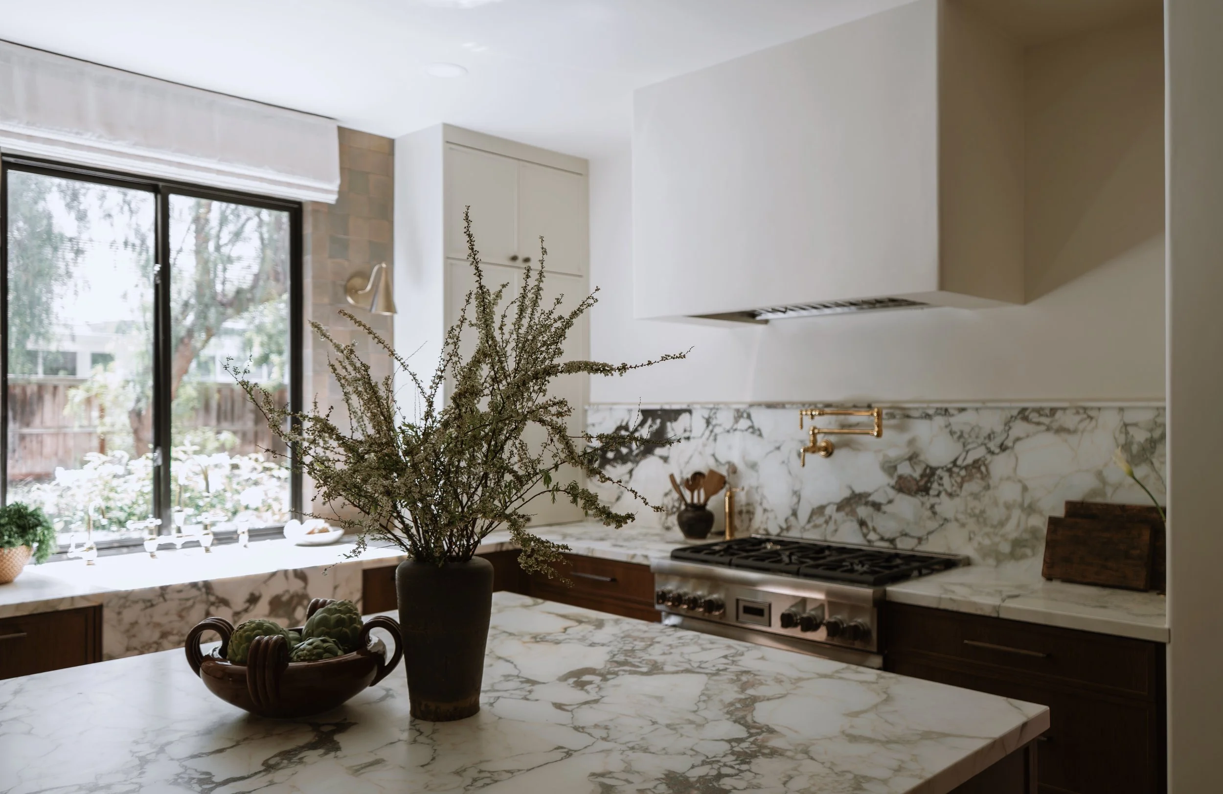 Modern kitchen with white cabinets, marble countertops, and a large window with a view of outside trees. Decor includes a vase with branches and a bowl of artichokes.