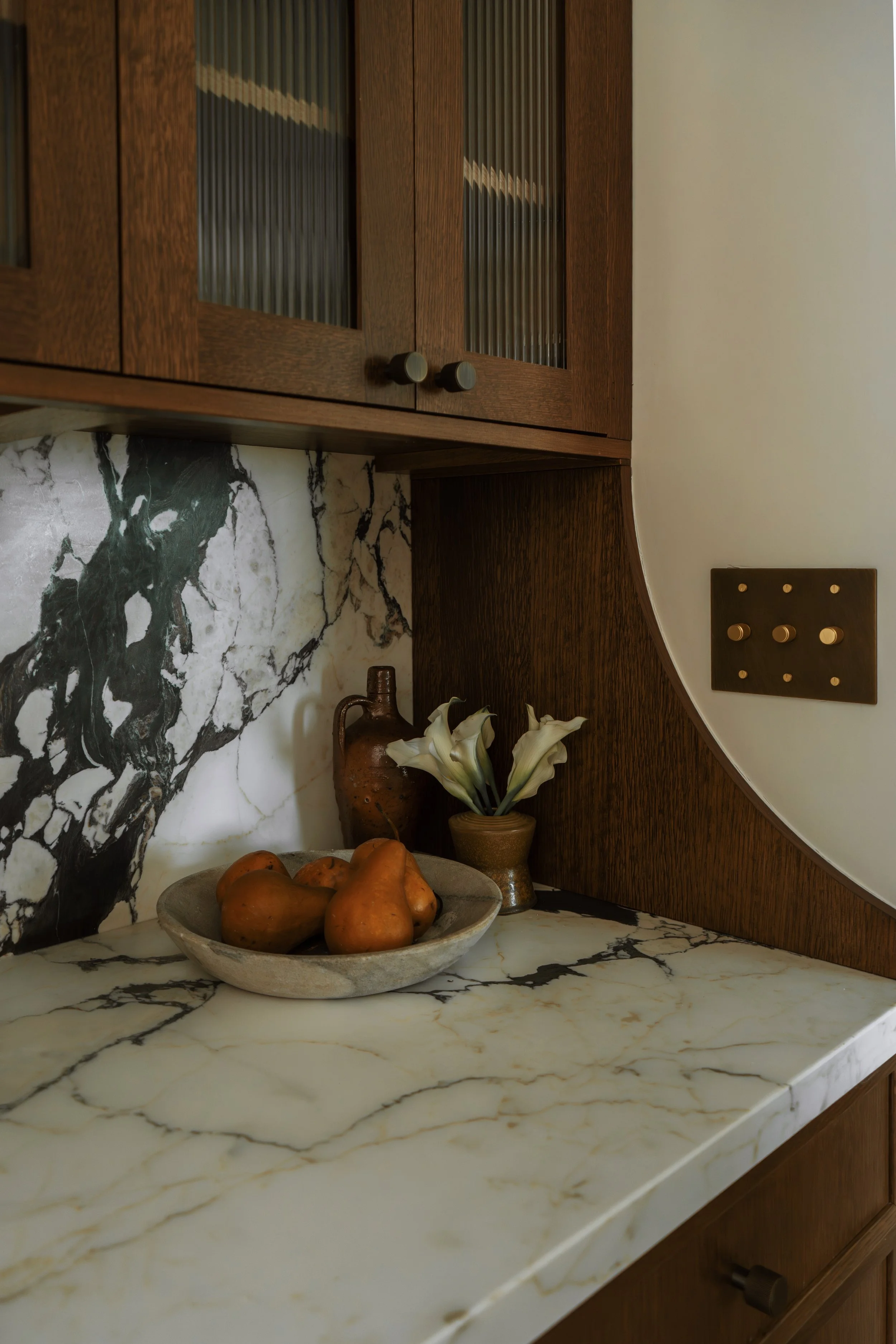 A kitchen countertop with a marble texture, a bowl of pears, a ceramic vase with white lilies, and a dark wooden cabinet with glass-paneled doors.