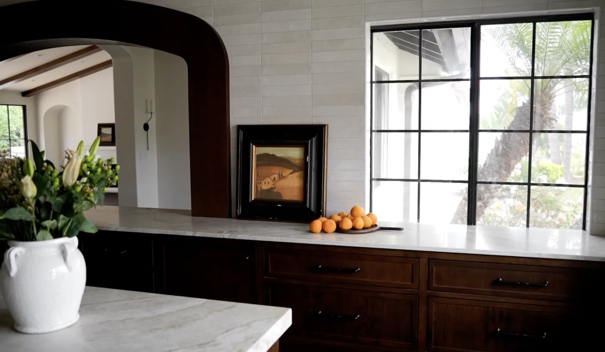 Kitchen with white marble countertop, dark wood cabinets, large window, and decorative items including a painting, a tray of oranges, and a vase of flowers.