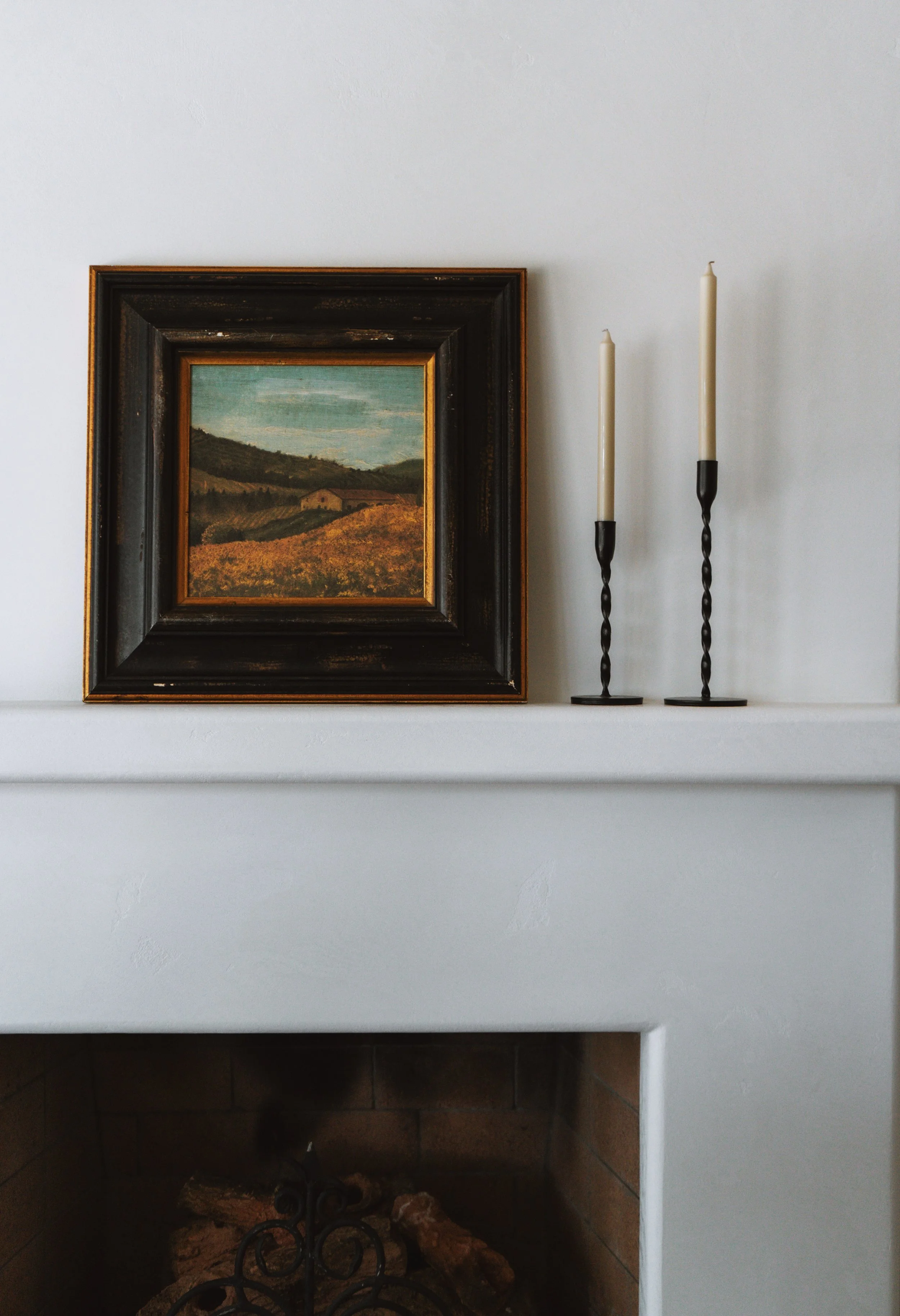 Interior of a white fireplace mantel with a landscape painting and two tall candlesticks with twisted black stands.