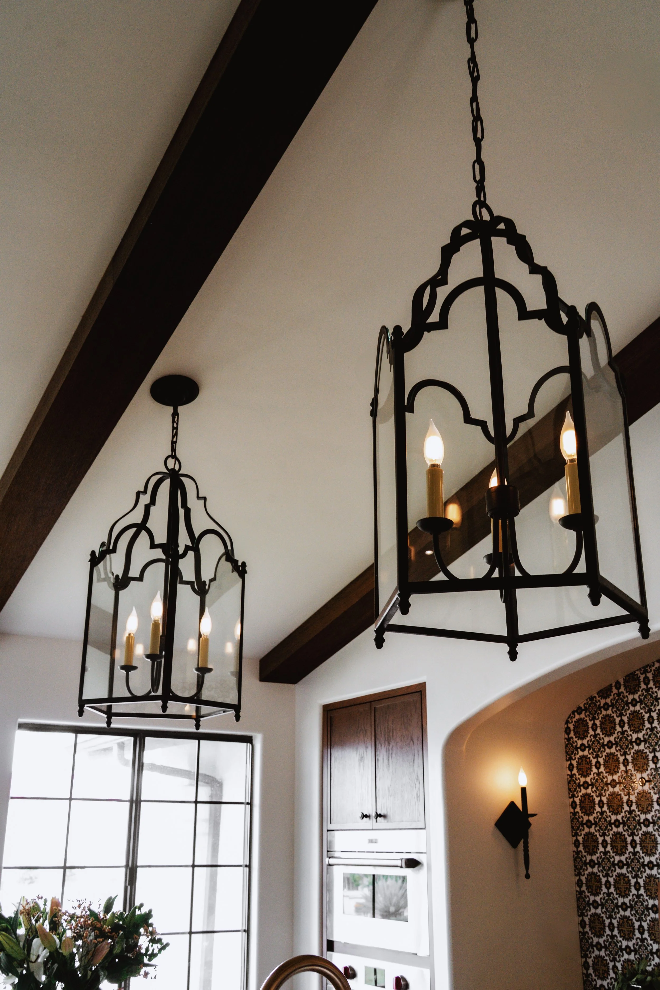 Two black metal chandeliers with candle-shaped light bulbs hanging from a white ceiling with wooden beams in a room with a large window and floral wallpaper.