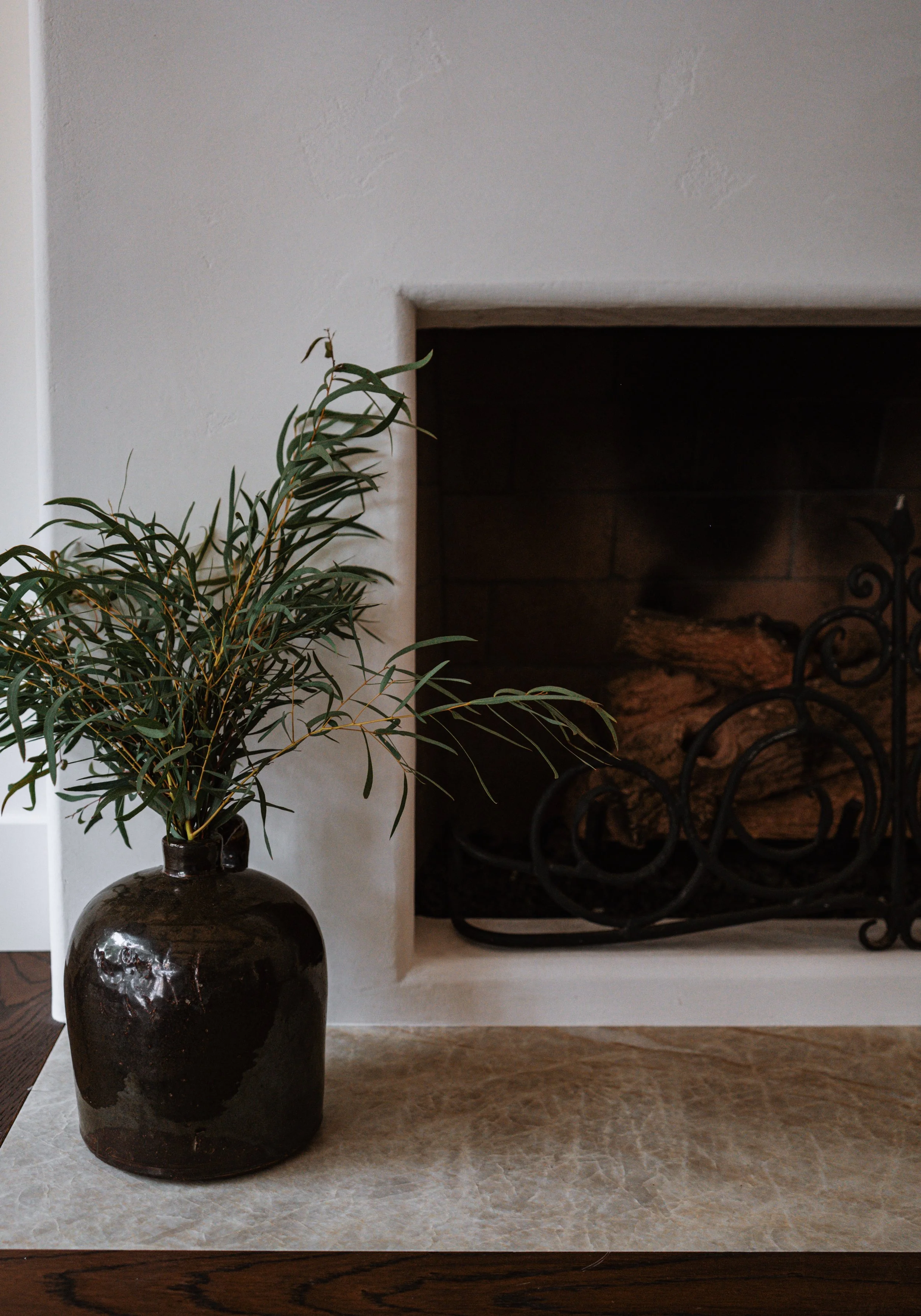 A brown ceramic vase with a glossy finish holding a leafy green plant placed on a white surface in front of a fireplace.