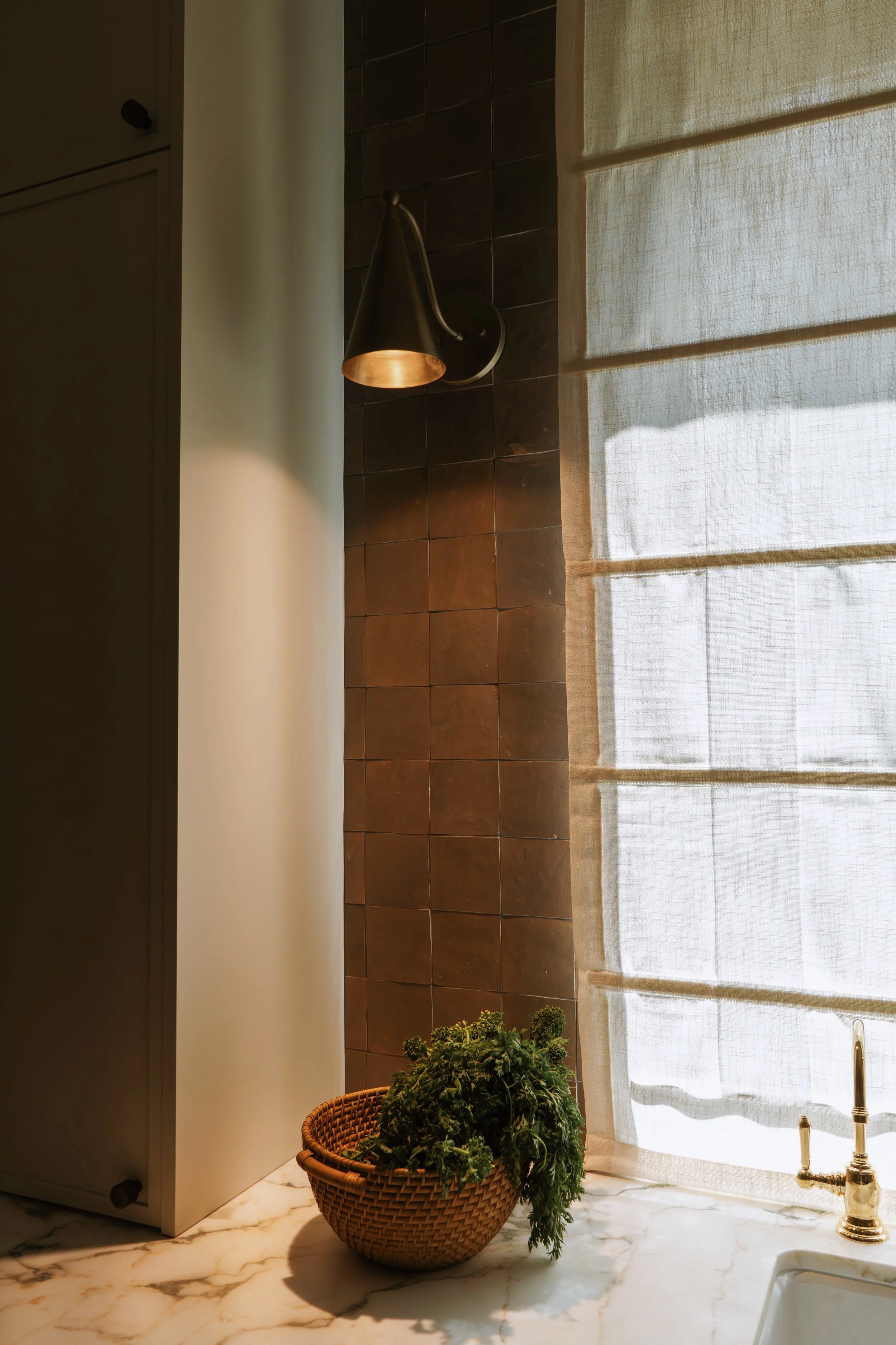 A kitchen countertop with a marble pattern, a basket with green herbs, a window with sheer curtains, a black wall-mounted lamp, and a sink faucet.