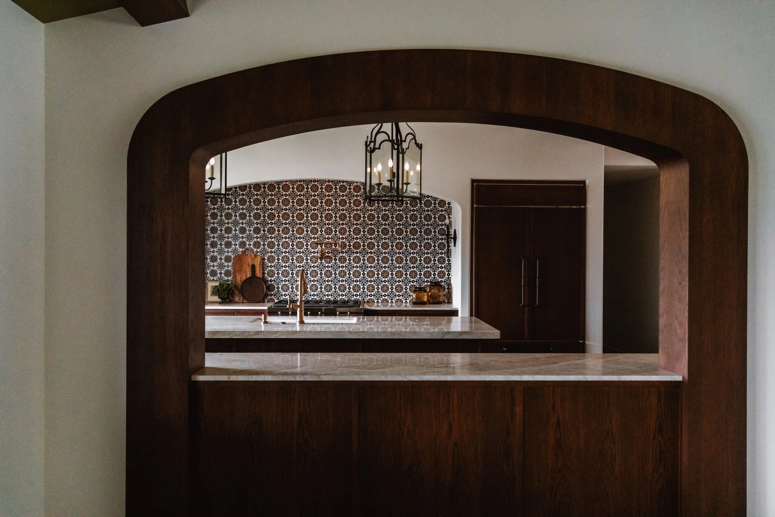 View through a large dark wood archway into a kitchen with patterned backsplash, hanging chandelier, and various kitchen items.