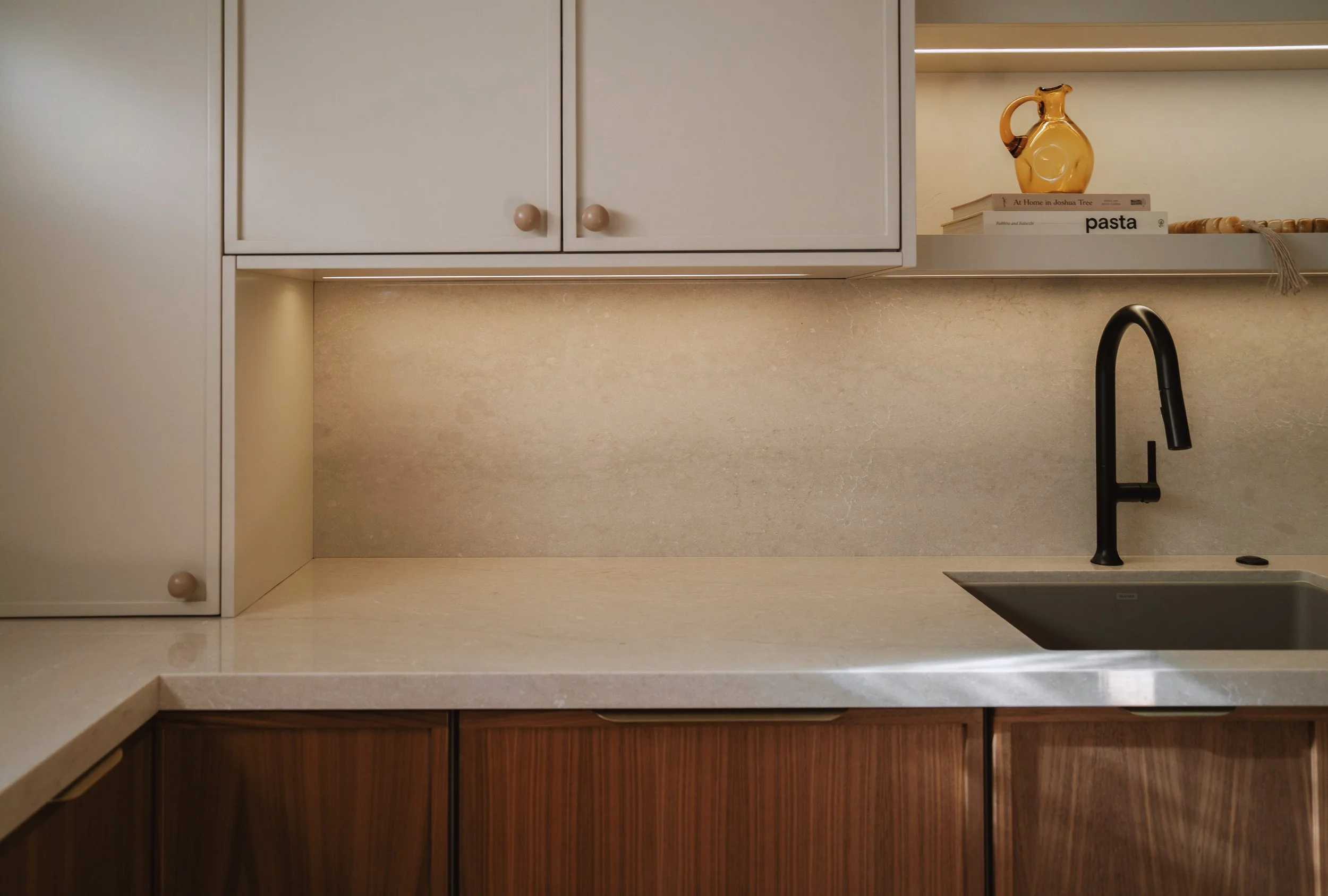 A modern kitchen countertop with a black faucet, beige backsplash, white cabinets, and a small open shelf holding a ceramic pitcher, a stack of books, and some decorative objects.