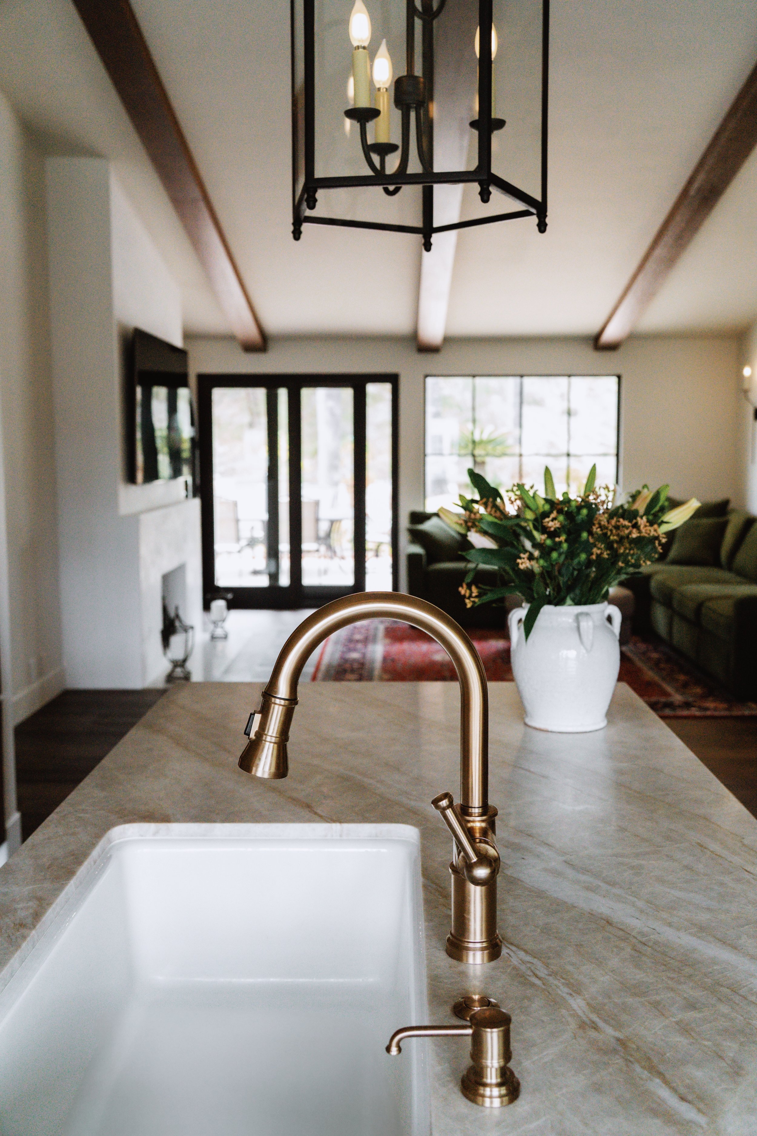 Close-up view of a kitchen island with a gold-colored faucet and a white sink, with a vase of flowers in the background. Living room with large windows, a sofa, and a dining table is visible beyond.