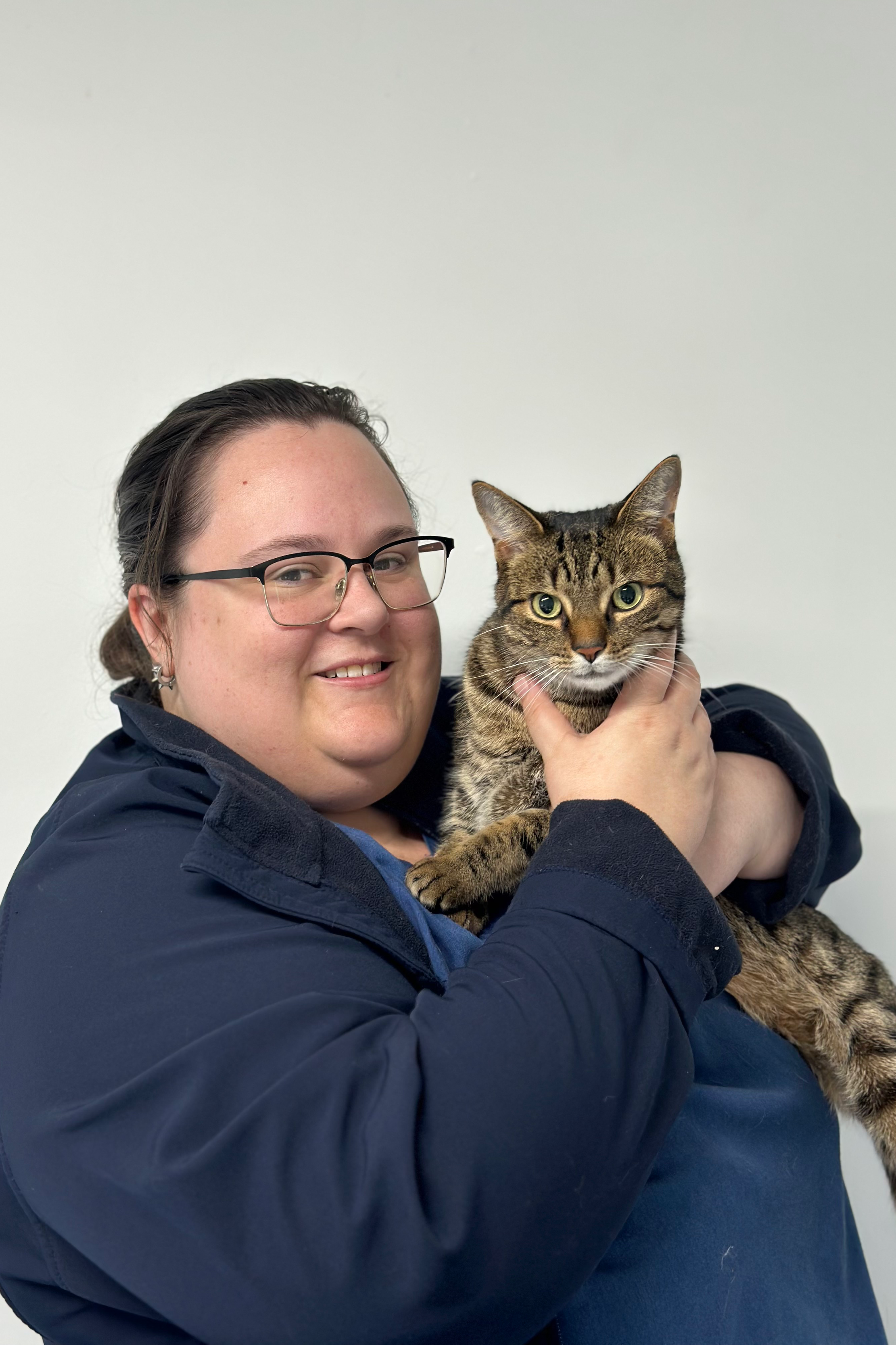 Person holding a tabby cat, wearing glasses and a navy jacket, against a plain background.