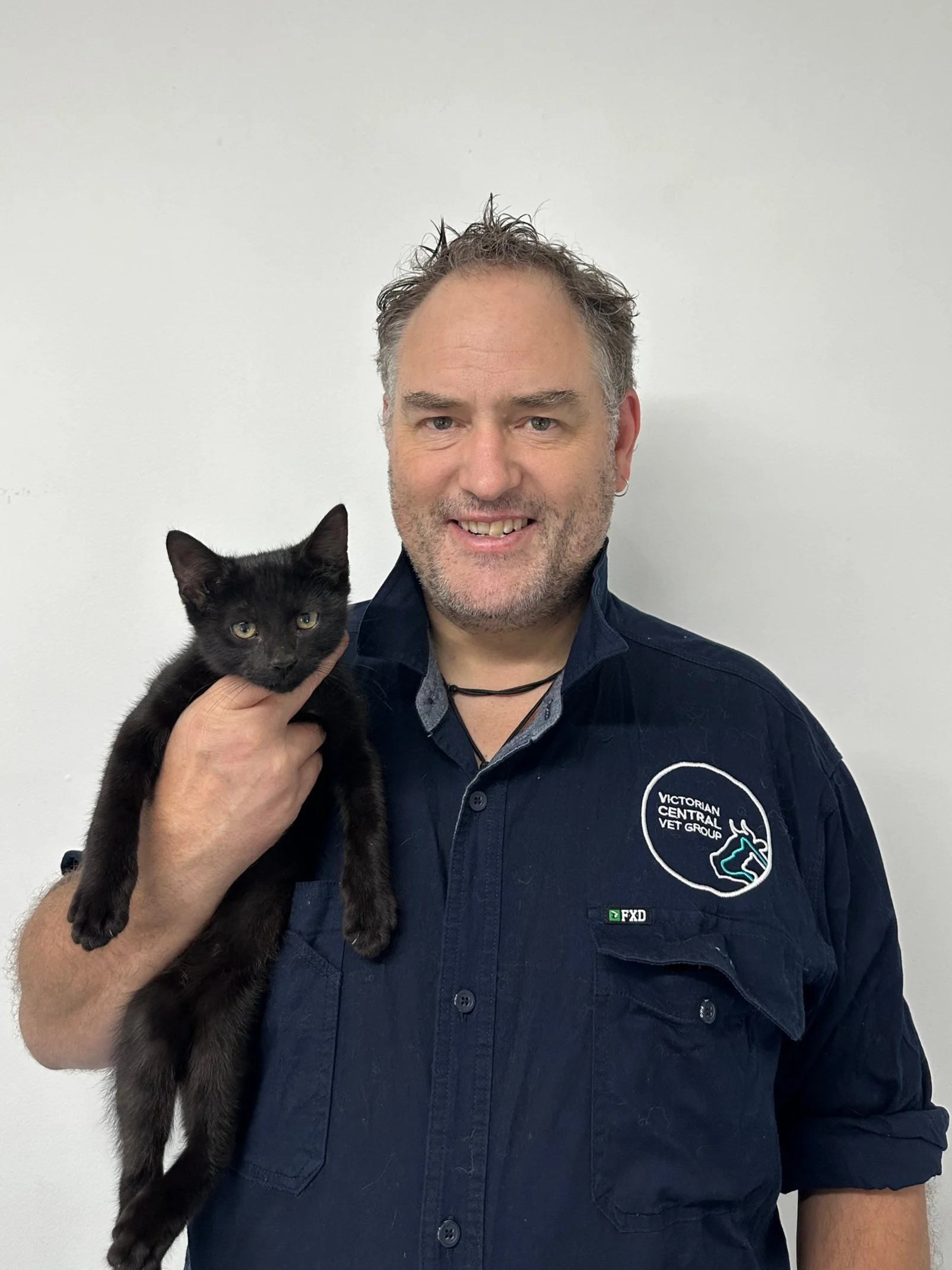 Man holding a black cat, smiling, wearing a dark blue shirt with a logo on it.