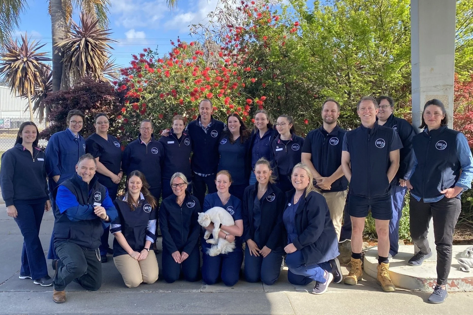 Group of people outdoors in uniform with jackets, posing with a white cat, in front of red flowering shrubs and palm trees.