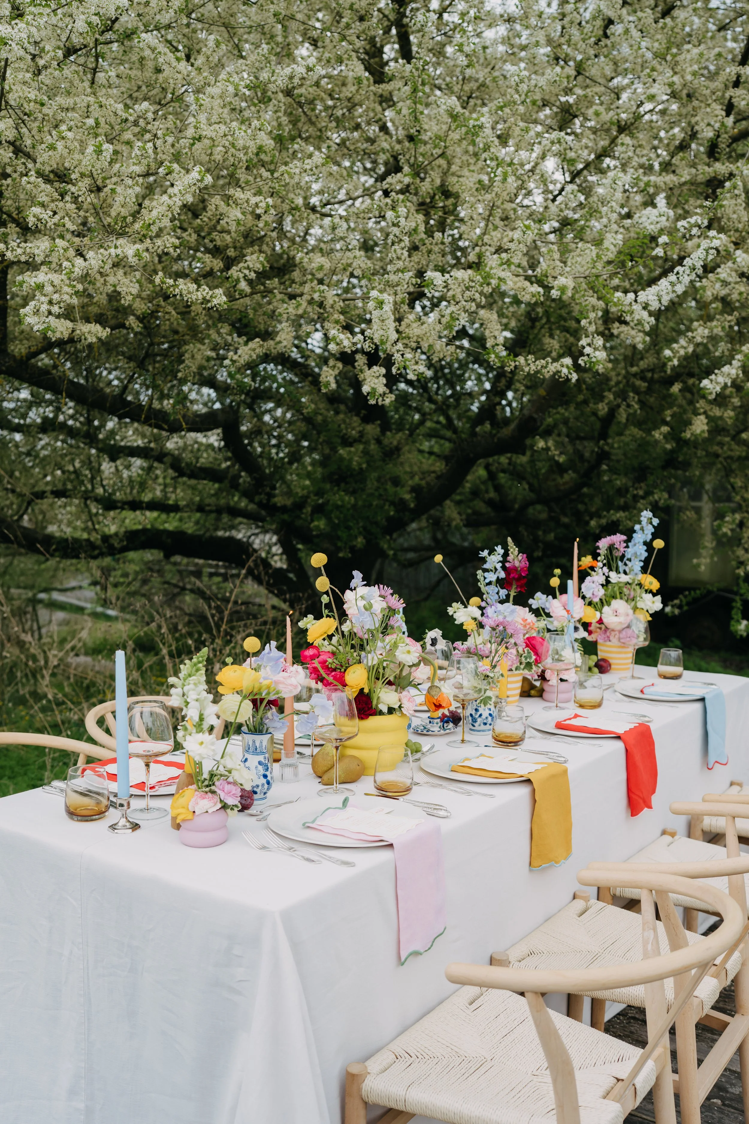 Bunter Outdoor-Tisch im Hofgarten der Blauen Halle – kreative Blumendeko in Gelb, Rosa und Blau unter blühendem Baum