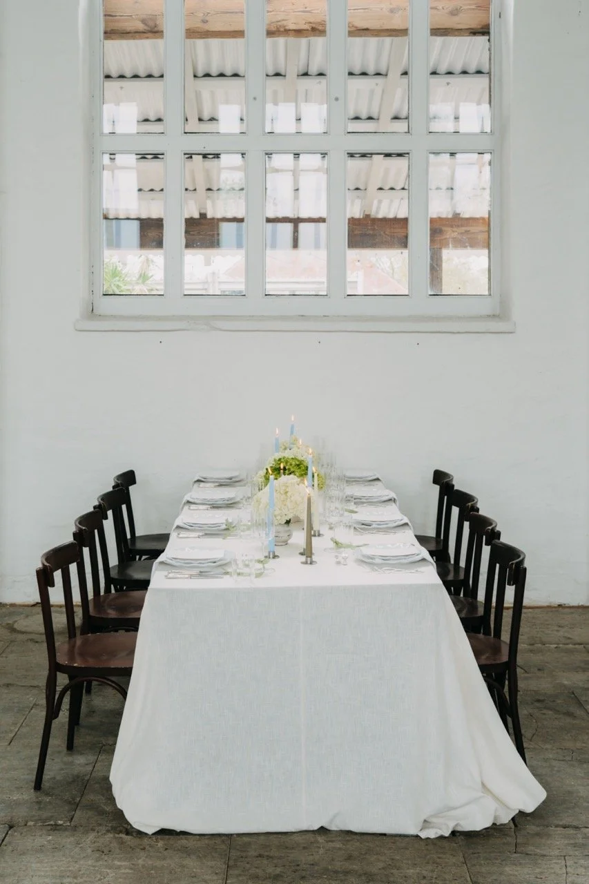Minimalistisch gedeckter Tisch in der Blauen Halle – Vintage Mobiliar, weiße Tischwäsche, Kerzen und moderne Eleganz vor weißer Wandkulisse