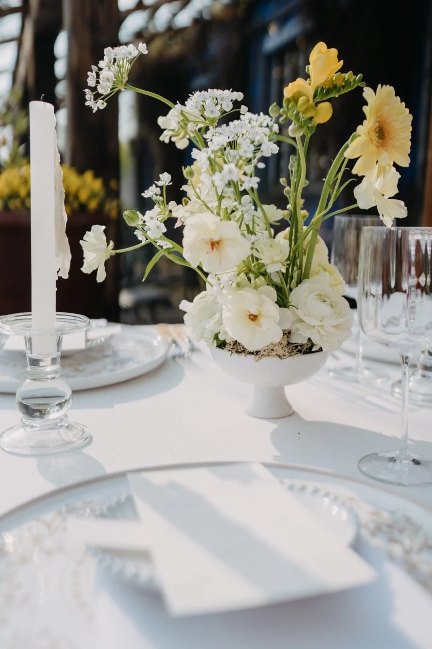 Detailaufnahme einer floralen Tischdekoration – zarte Creme- und Gelbtöne mit Freesien, Ranunkeln und Schleierkraut in Fußschale auf weiß eingedecktem Hochzeitstisch bei der Blauen Halle