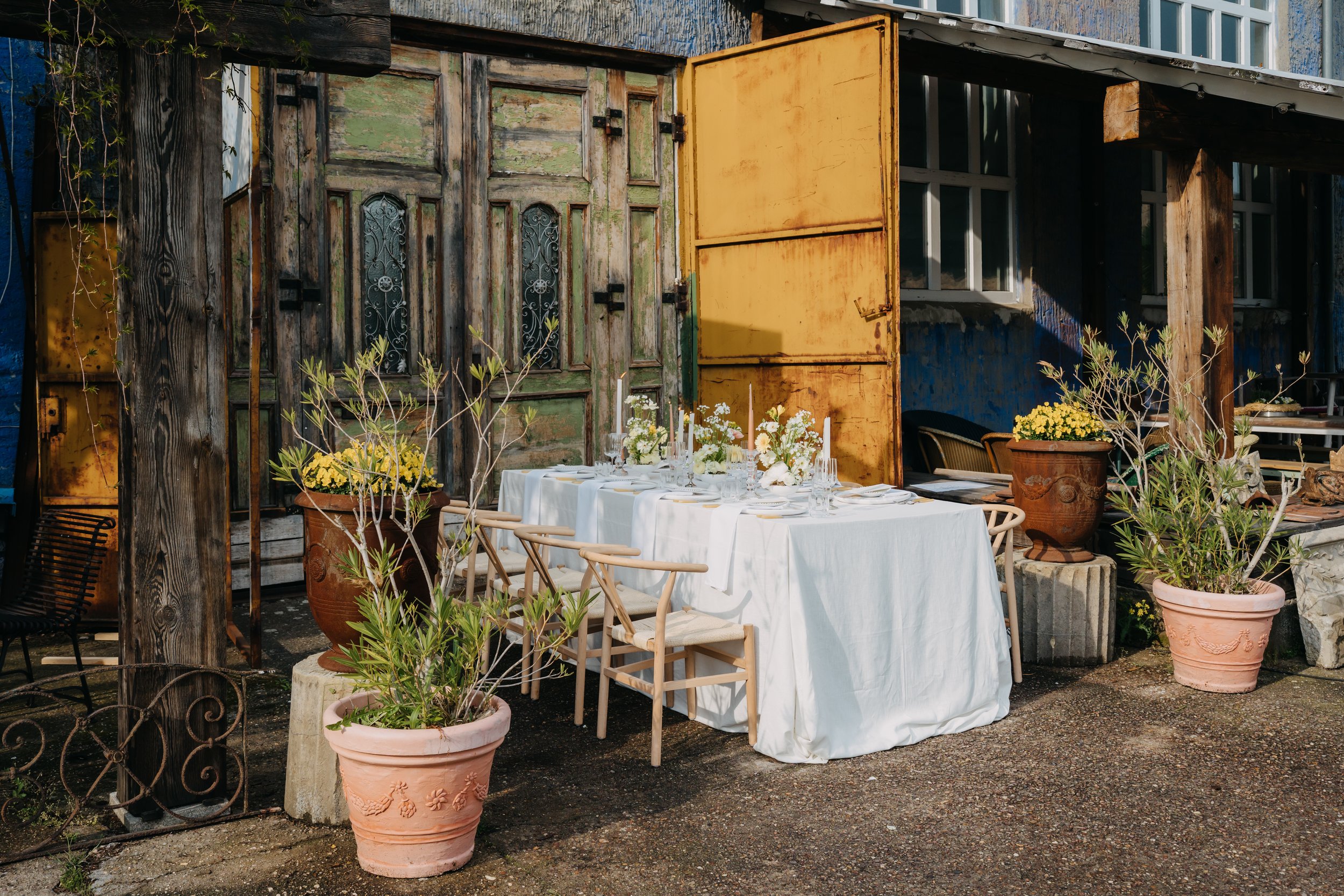 Outdoor-Dinnertisch vor der Blauen Halle, rustikale Holztüren, mediterrane Pflanzkübel und weiß eingedeckte Festtafel für exklusive Events.

